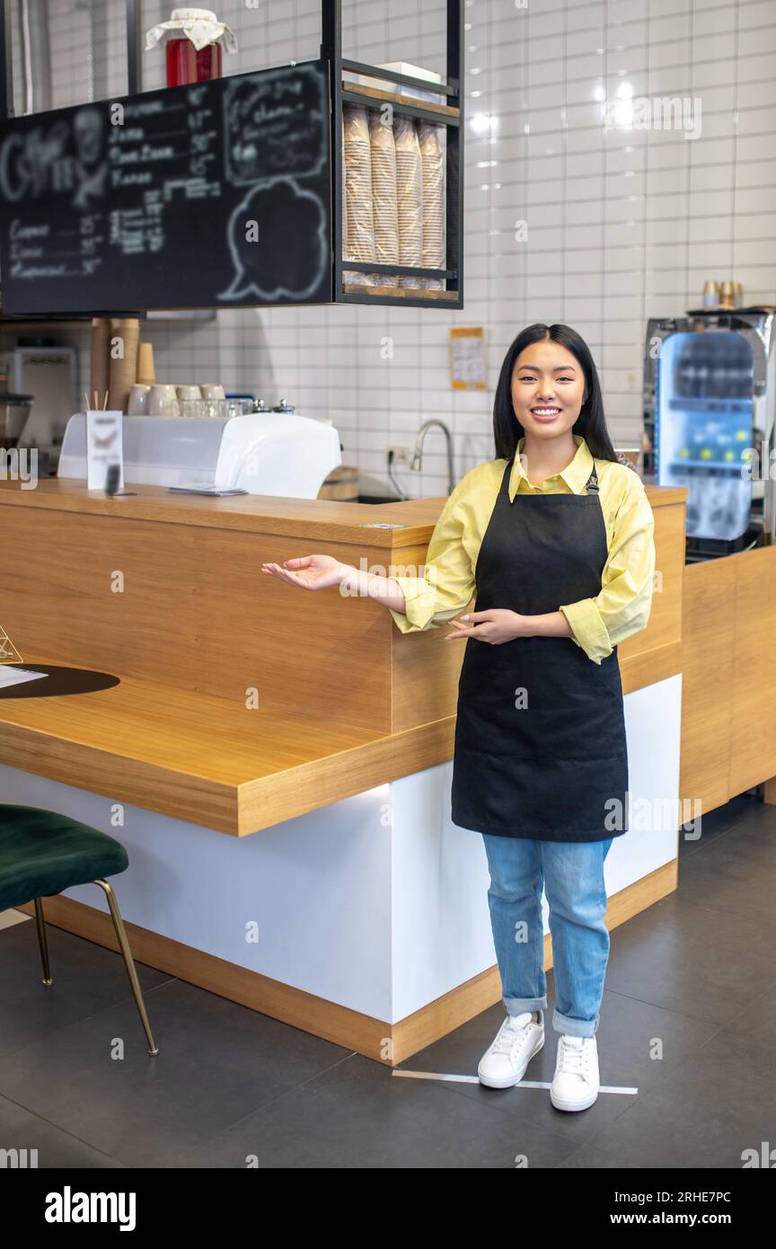 Mignonne serveuse souriante dans un tablier noir debout près du comptoir du café Banque D'Images