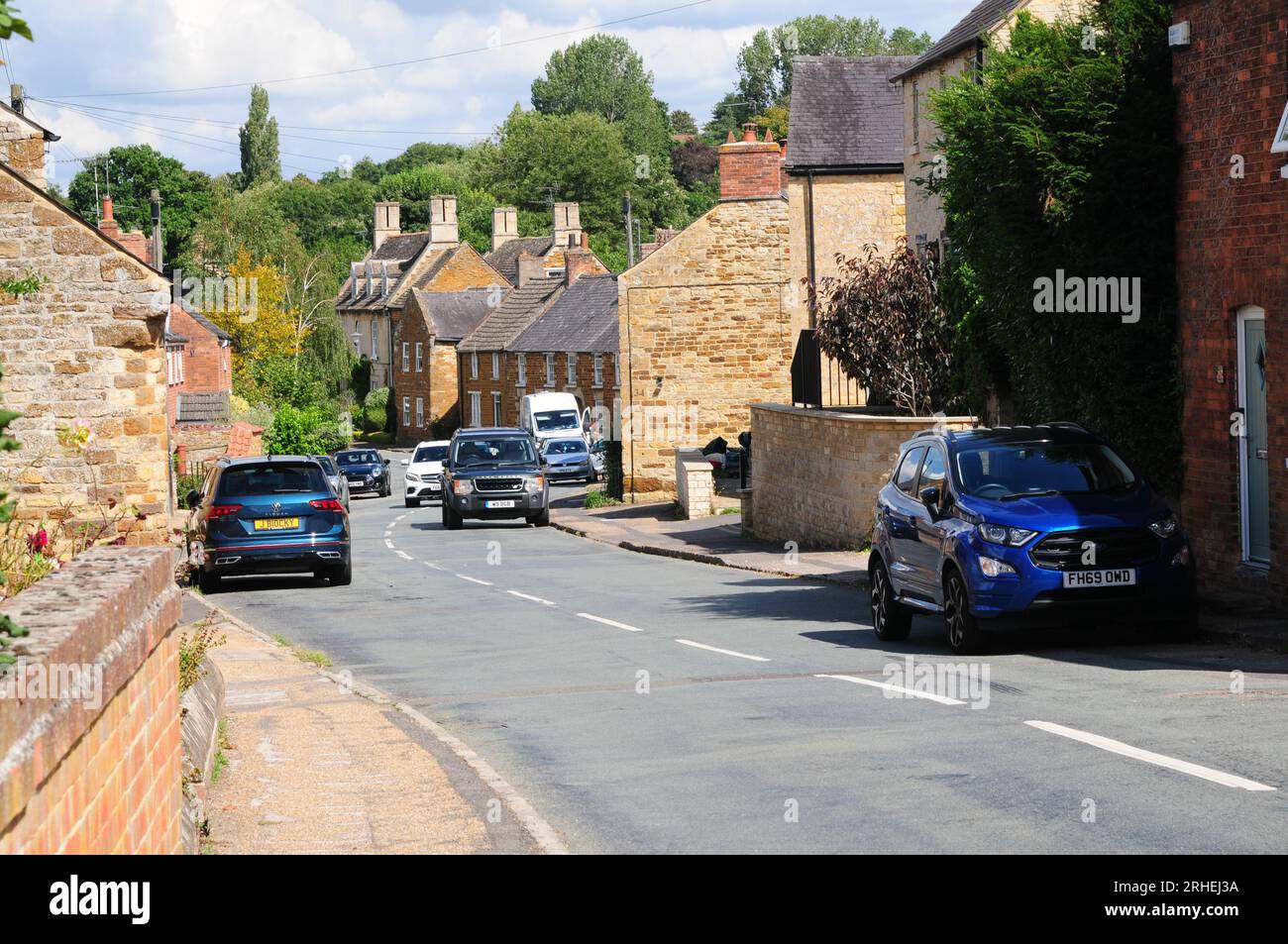 Scène de rue de village avec des cottages en pierre de fer, des voitures garées et la circulation Banque D'Images