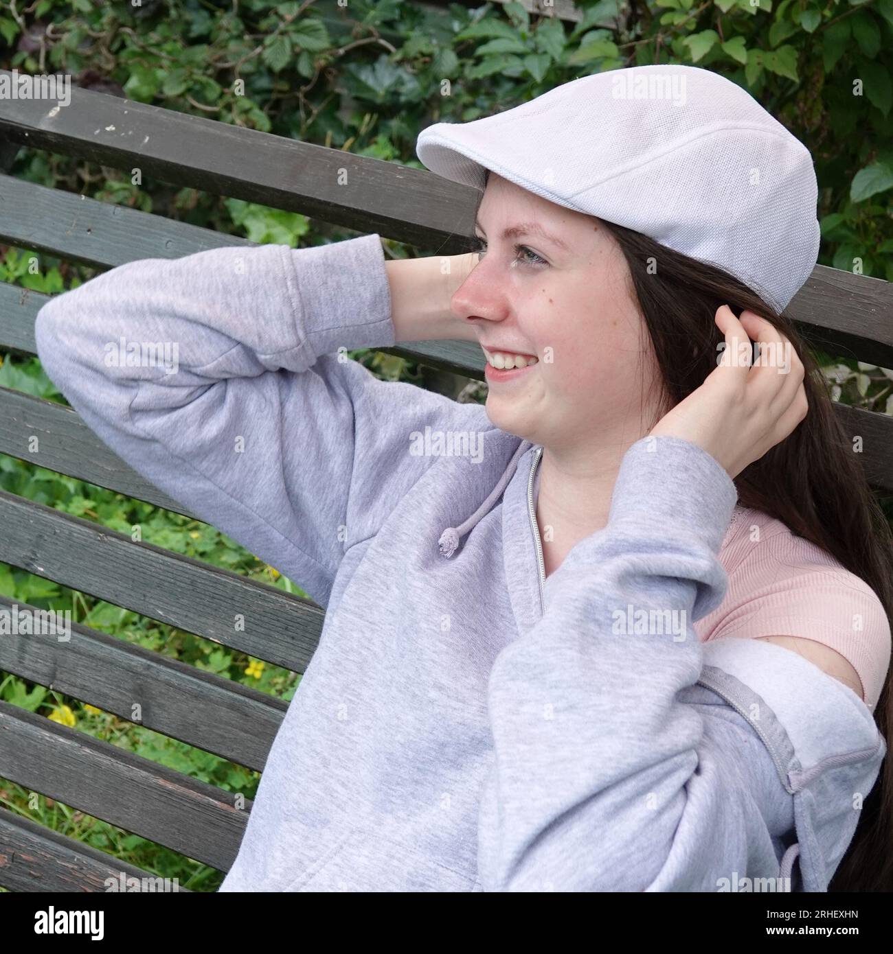 Une adolescente avec une casquette paddy blanche s'assoit en souriant large et touche ses cheveux brun foncé. Banque D'Images