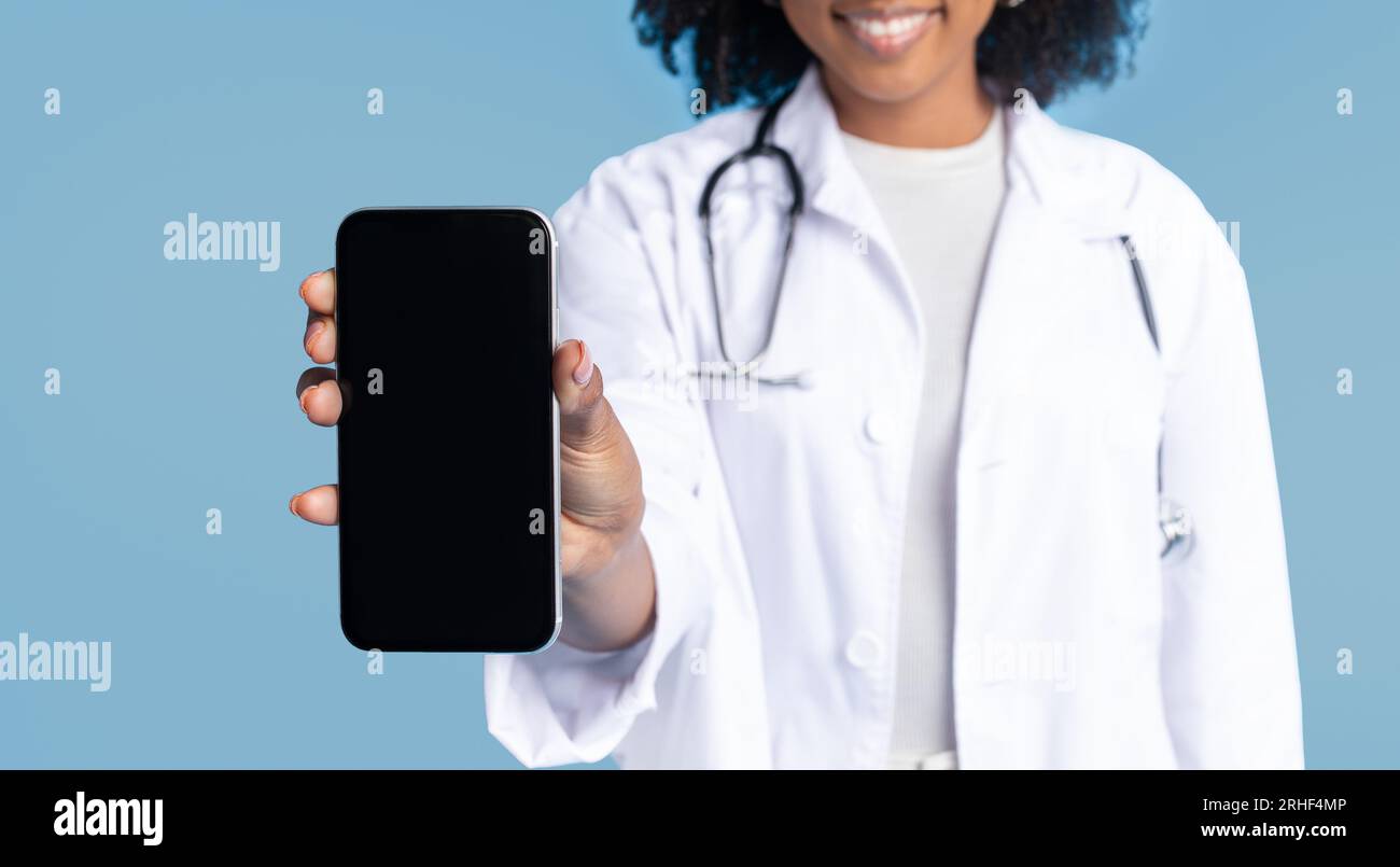 Souriant noir millennial dame médecin en manteau blanc montrer smartphone avec écran vide Banque D'Images