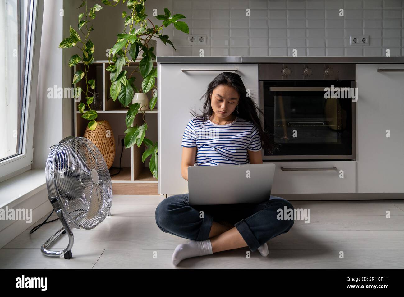 Beau jeune fille asiatique étudiante avec ordinateur portable languissant la chaleur assis sur le plancher frais à côté du ventilateur. Banque D'Images