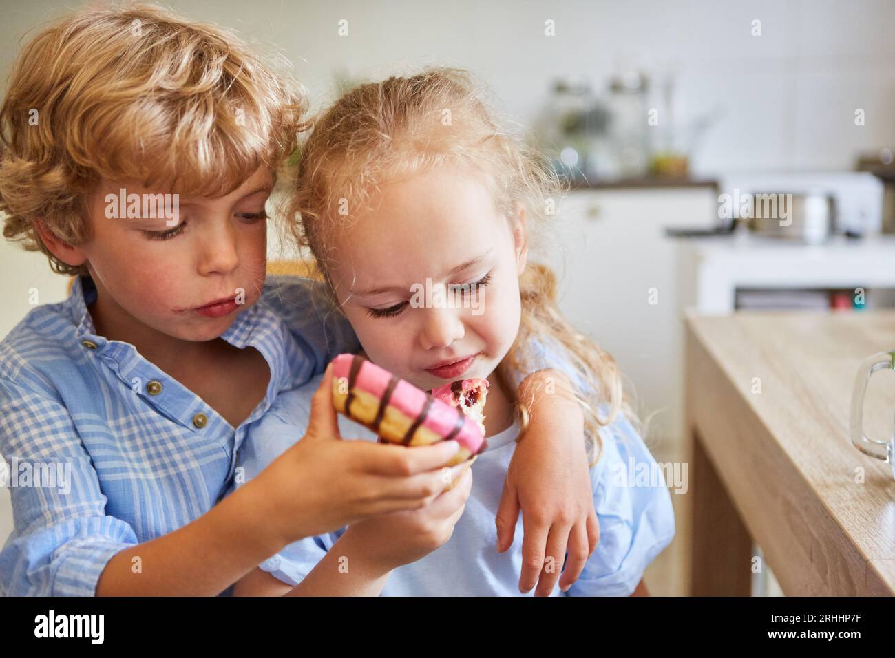 Garçon blond partageant des beignets avec sa sœur près de la table à la maison Banque D'Images Garçon blond partageant des beignets avec sa sœur près de la table à la maison Banque D'Images