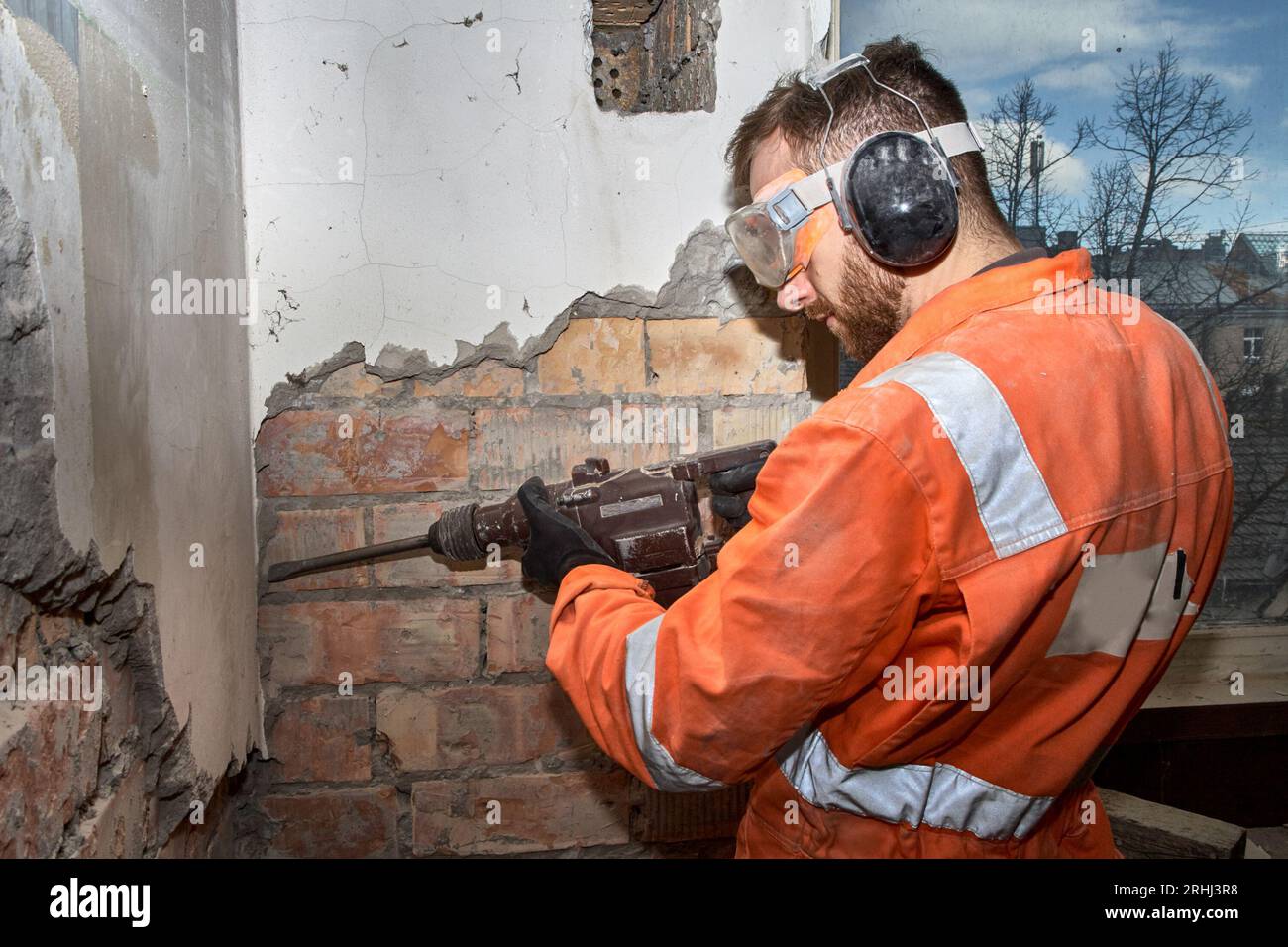 Travailleur de la construction utilisant une perceuse à percussion avec burin pour les travaux de démolition à l'intérieur, portant une combinaison orange et un EPI. Banque D'Images