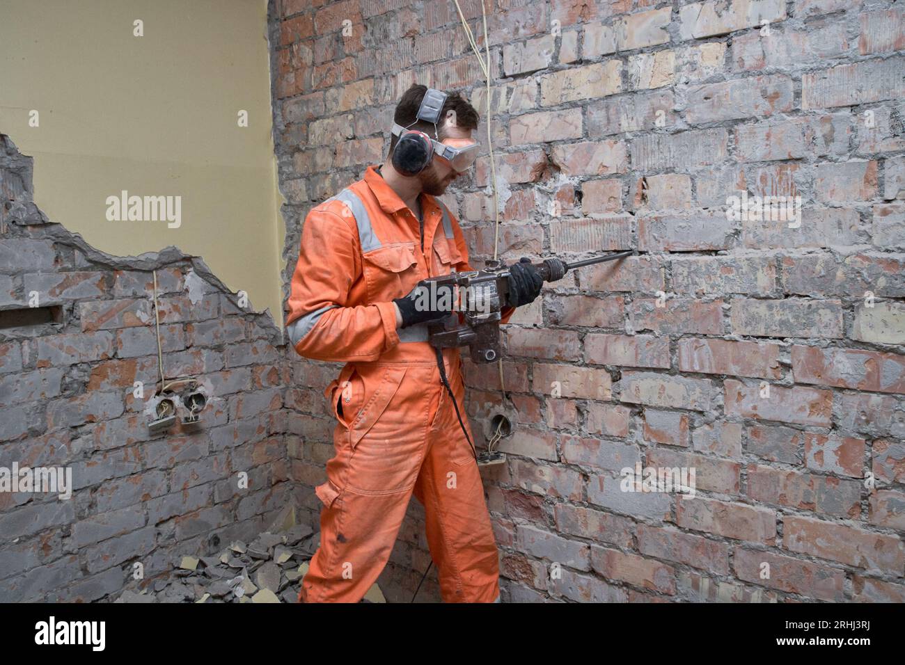 Travailleur de la construction à l'aide d'une perceuse à percussion avec burin pour enlever le vieux ciment du mur de briques à l'intérieur, portant des gants, un masque, des casques antibruit et orange covera Banque D'Images