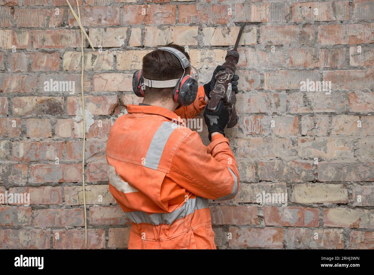 Travailleur de la construction enlevant le vieux plâtre du mur de briques rouges à la perceuse à percussion en position verticale, portant des gants, un masque, un casque antibruit et une couverture orange Banque D'Images