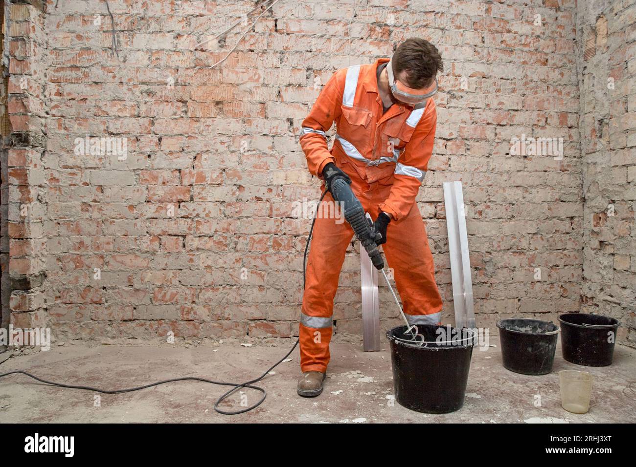 Constructeur utilisant le mélangeur pour pétrir la solution de ciment dans le seau. Travailleur portant un EPI et une combinaison orange. Banque D'Images