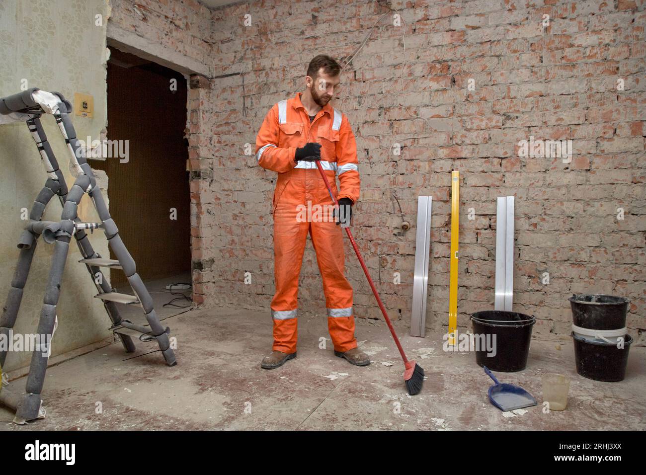 Travailleur de la construction homme nettoyant la salle avec un balai après les travaux de démolition, portant des gants, des bottes et des combinaisons orange. Banque D'Images