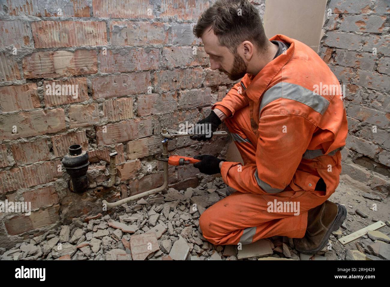 Jeune plombier mâle fixant le raccord d'eau qui fuit, portant des gants, des bottes et une combinaison orange. Banque D'Images