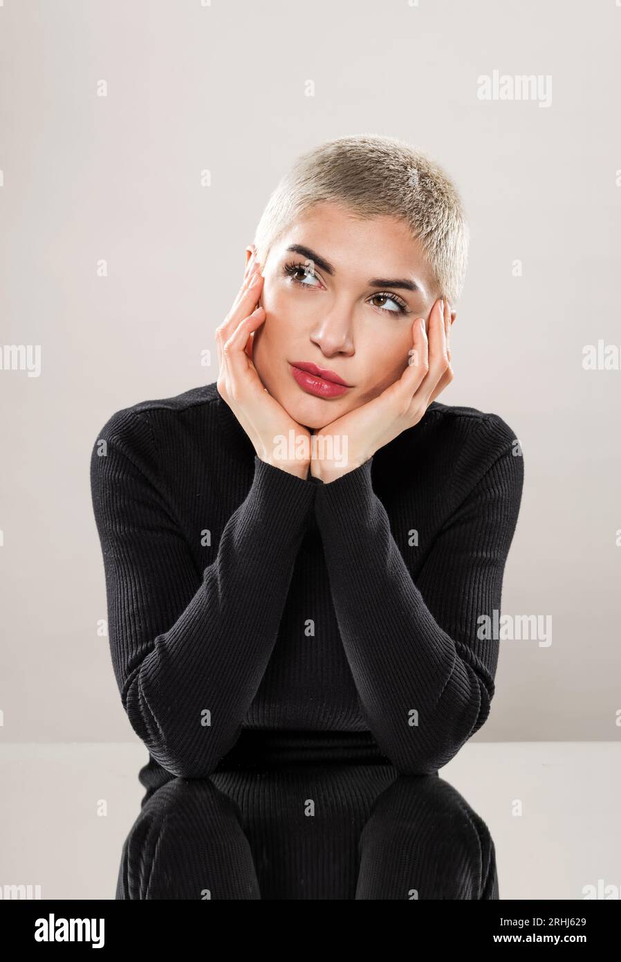 Charmante jeune femme aux cheveux rasés courts portant une chemise noire appuyée sur les mains tout en rêvant et regardant loin dans le studio Banque D'Images