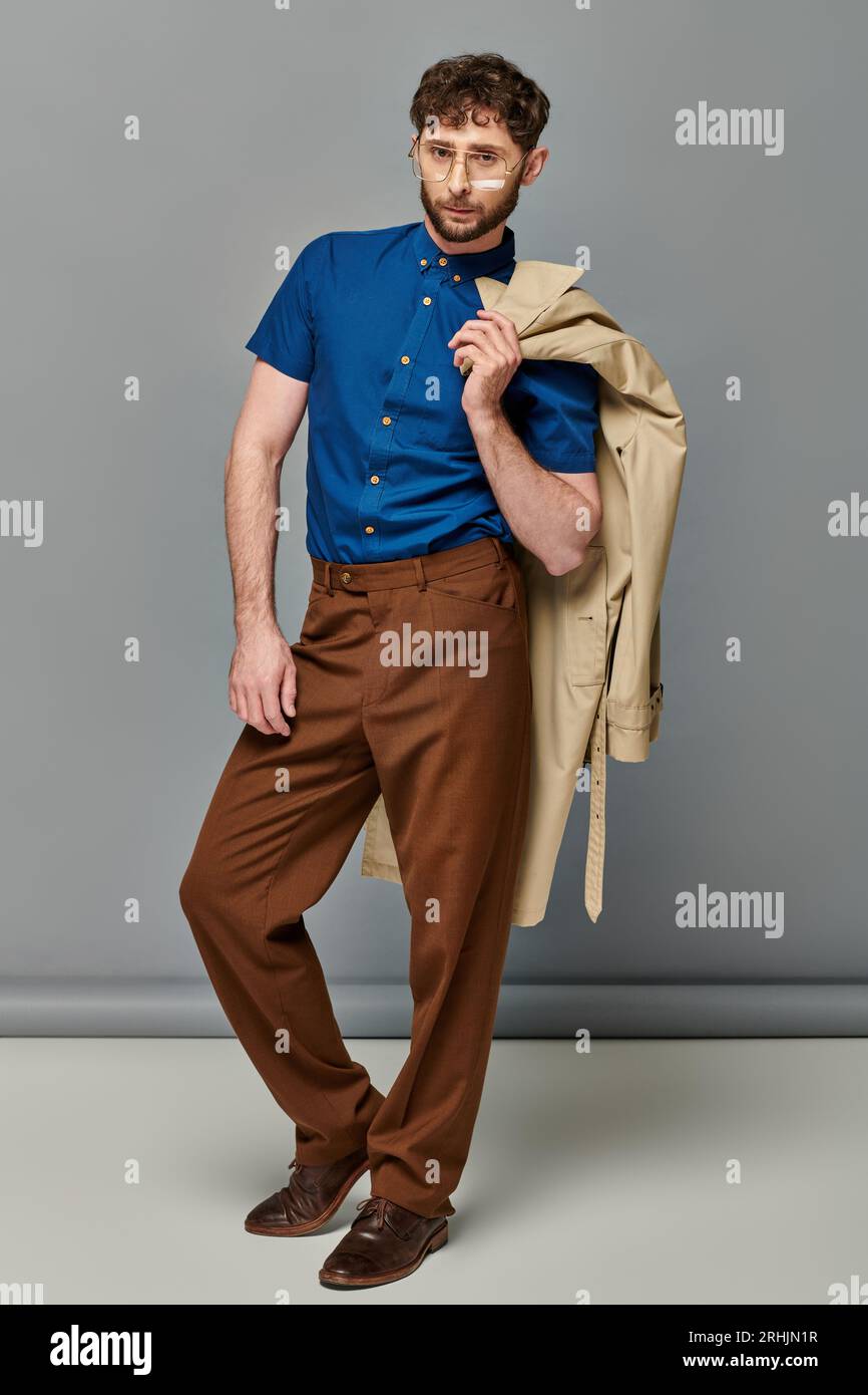 homme barbu élégant et décontracté dans des lunettes tenant un trench-coat, fond gris, modèle masculin à la mode Banque D'Images