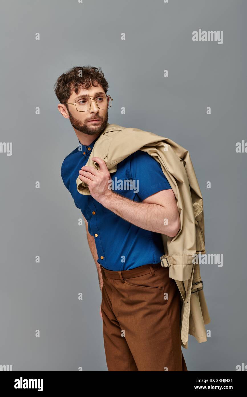 homme barbu dans des lunettes tenant le trench coat, fond gris, modèle masculin regardant loin, chic décontracté Banque D'Images