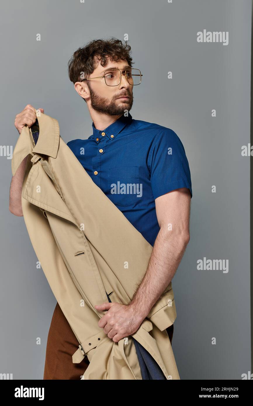 homme dans les lunettes tenant le trench coat, fond gris, modèle masculin élégant regardant loin, chic décontracté Banque D'Images