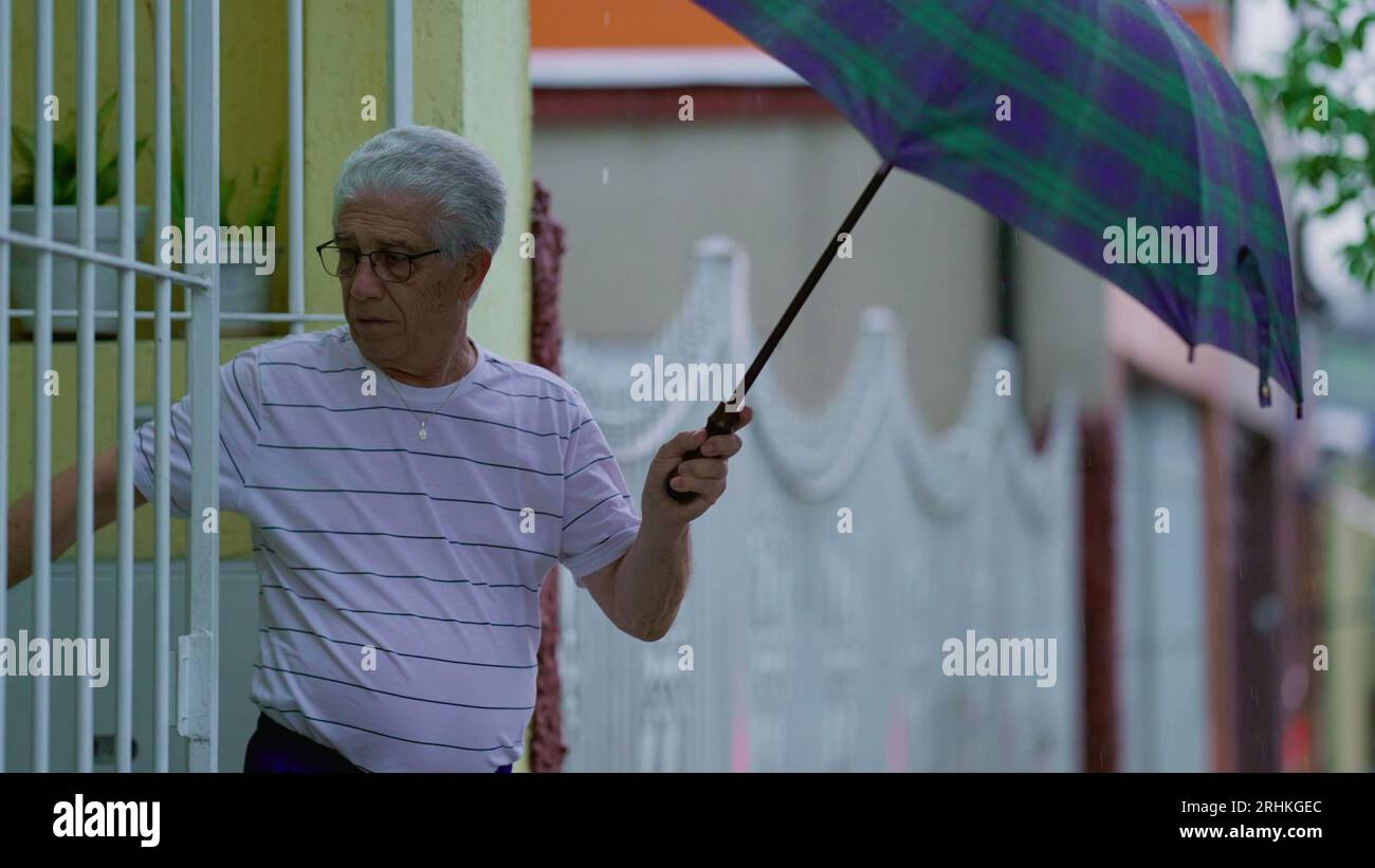 Homme senior ouvrant un parapluie, sort dans la rue sous la pluie, ferme la porte de la maison derrière lui. Personne âgée quittant sa résidence le jour de pluie à Slow-M. Banque D'Images