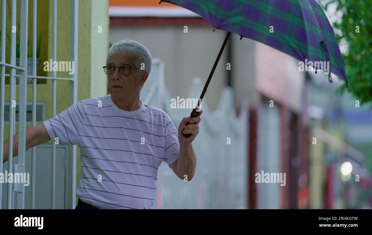 Homme senior ouvrant un parapluie, sort dans la rue sous la pluie, ferme la porte de la maison derrière lui. Personne âgée quittant sa résidence le jour de pluie à Slow-M. Banque D'Images