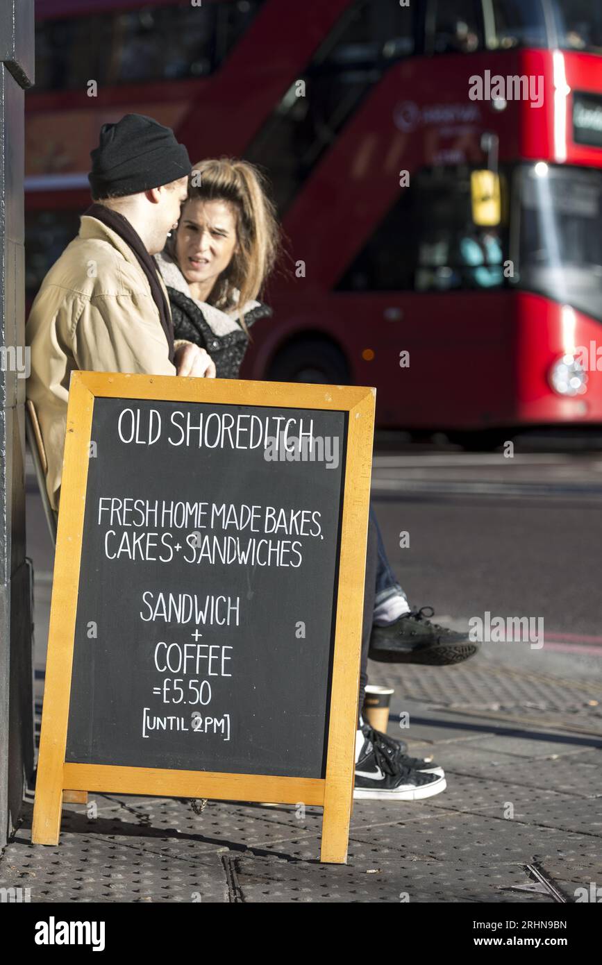 Couple à l'extérieur du café Old Shoreditch avec menu tableau noir et bus rouge de Londres. Remarque : le café a depuis fermé. Usage éditorial exclusif. Banque D'Images