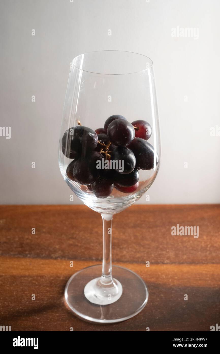 Un bouquet de raisins rouge foncé dans un verre à vin maintenu sur une table en bois sur fond blanc Banque D'Images