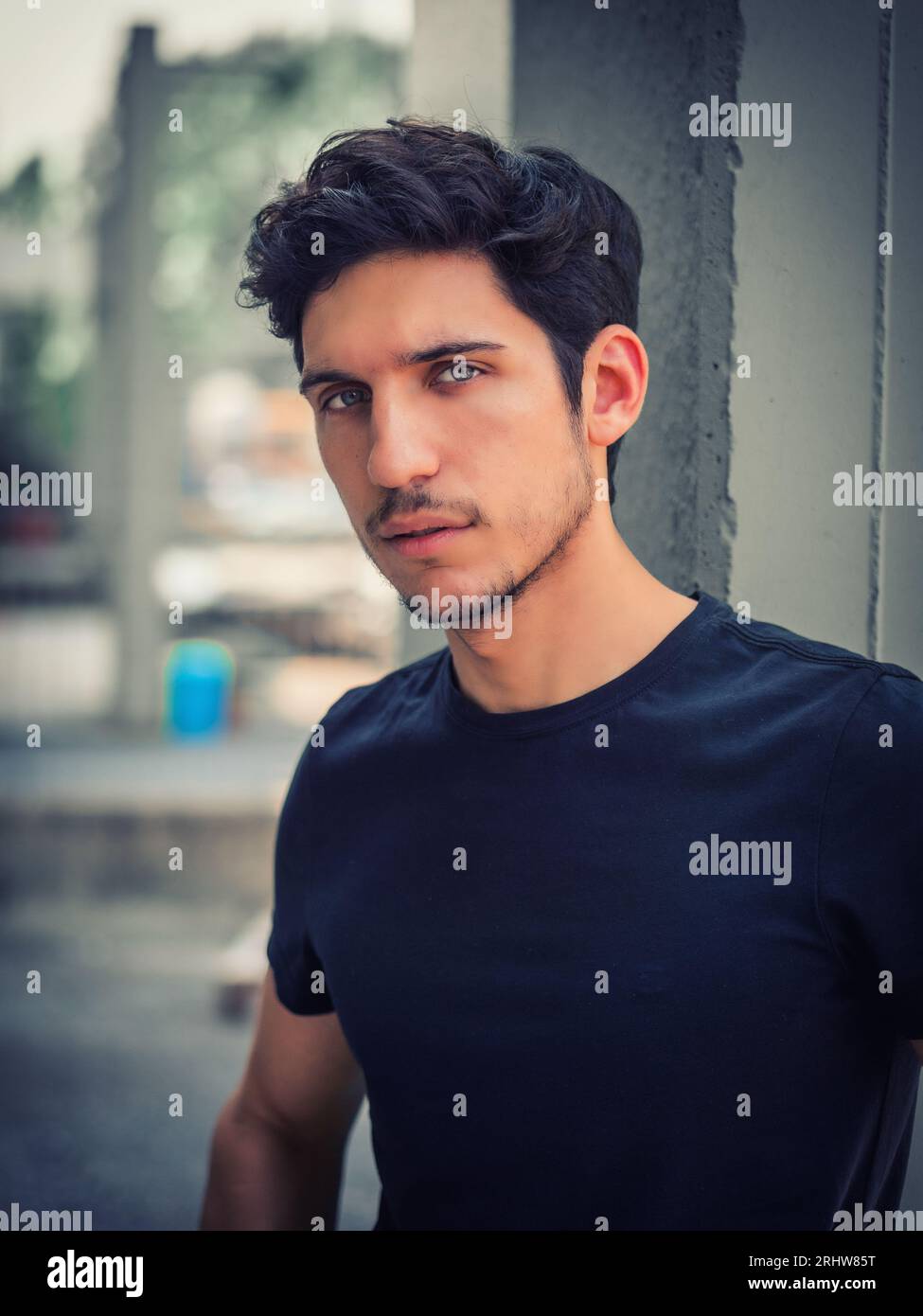 Photo de la tête et des épaules d'un beau jeune homme aux yeux verts, en milieu urbain appuyé contre un mur, regardant la caméra, portant un t-shirt Banque D'Images