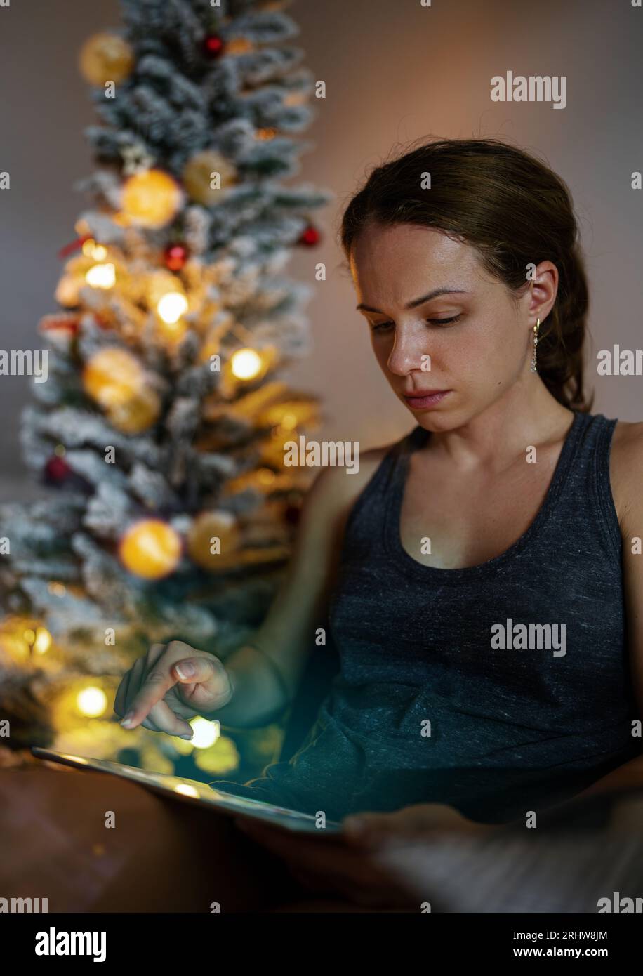 Brunette femme est assise dans la chaise à la maison pendant les vacances de Noël et la lecture ebook, sur le fond de l'arbre de Noël. Banque D'Images