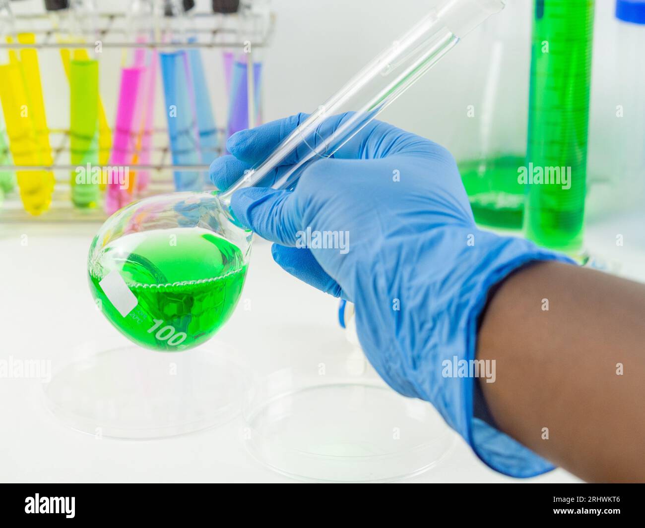 Gros plan d'une main portant des gants tenant une fiole jaugée avec un échantillon liquide vert dans un laboratoire. Banque D'Images