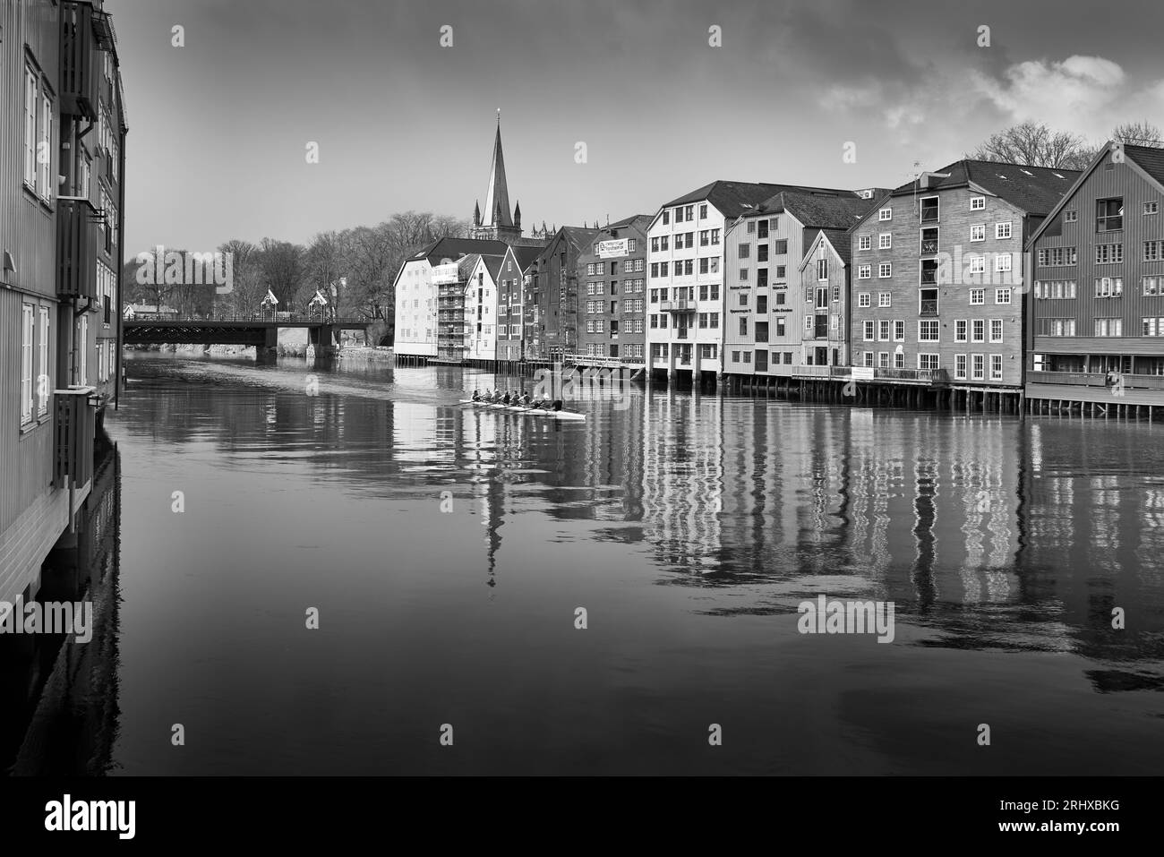 Photo en noir et blanc d'Un bateau aviron « Coxed Eight » passant devant des entrepôts riverains restaurés sur la rivière Nidelva, Bakklandet, Trondheim, Norvège Banque D'Images