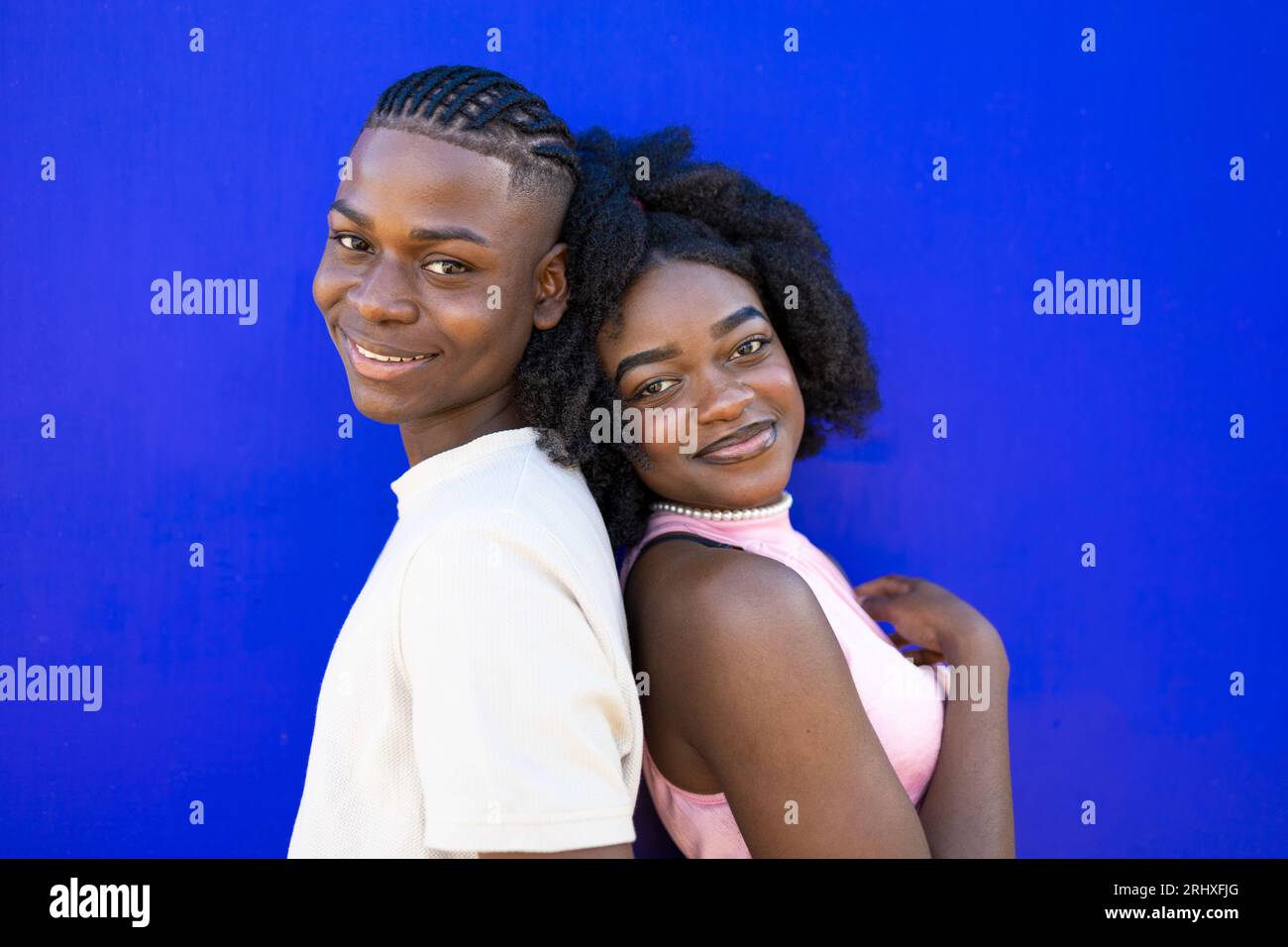 Portrait vue latérale de couple noir positif avec coiffure afro dans des vêtements décontractés souriant et regardant la caméra tout en se penchant dos à dos contre bleu b Banque D'Images