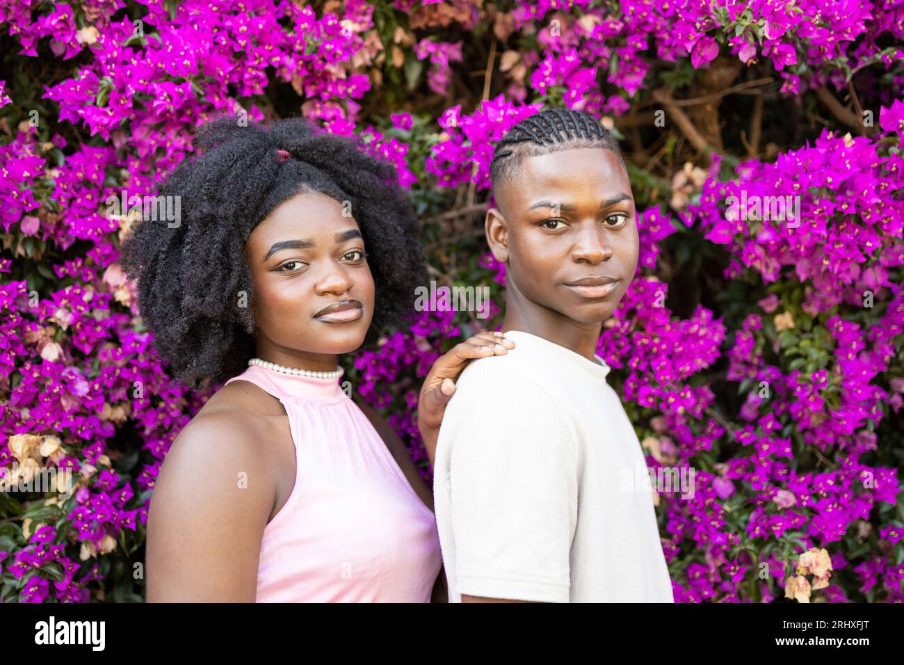 Vue de côté d'un couple adolescent afro-américain avec coiffure afro dans des vêtements décontractés tout en se tenant ensemble regardant la caméra dans le parc avec floraison pur Banque D'Images