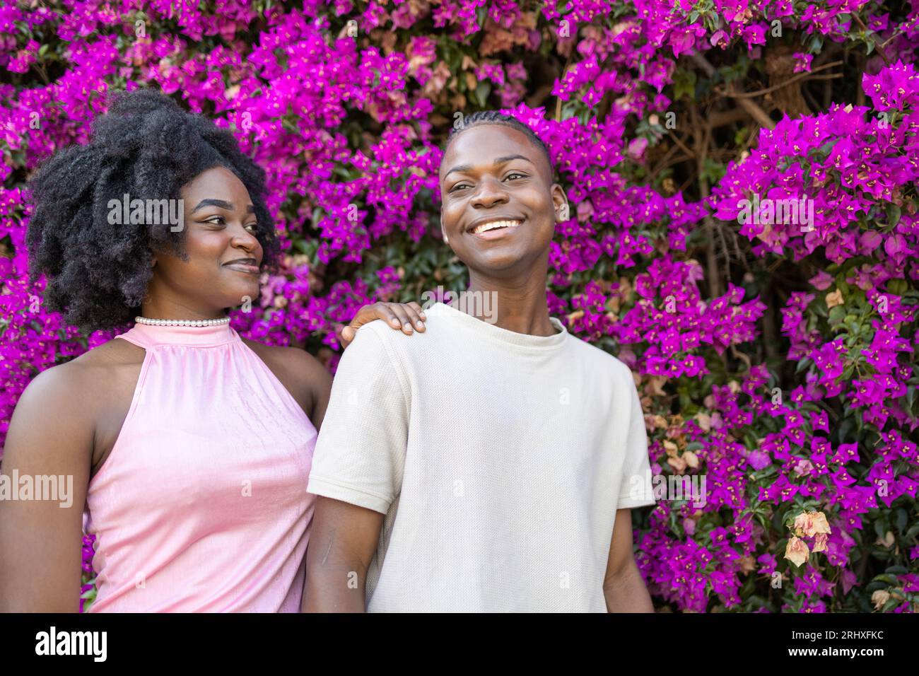 Couple d'adolescents afro-américains positif avec coiffure afro dans des vêtements décontractés souriant tout en se tenant ensemble dans le parc avec des fleurs violettes en fleurs Banque D'Images