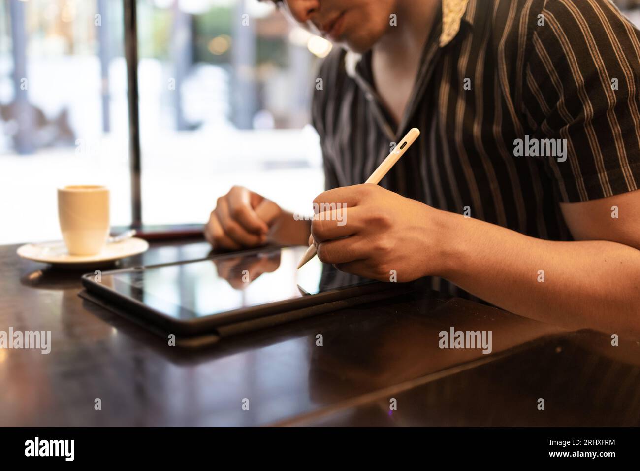 Anonyme beau jeune designer masculin dessinant sur tablette graphique avec stylo numérisé et prenant le café tout en étant assis par la fenêtre dans les cafés Banque D'Images