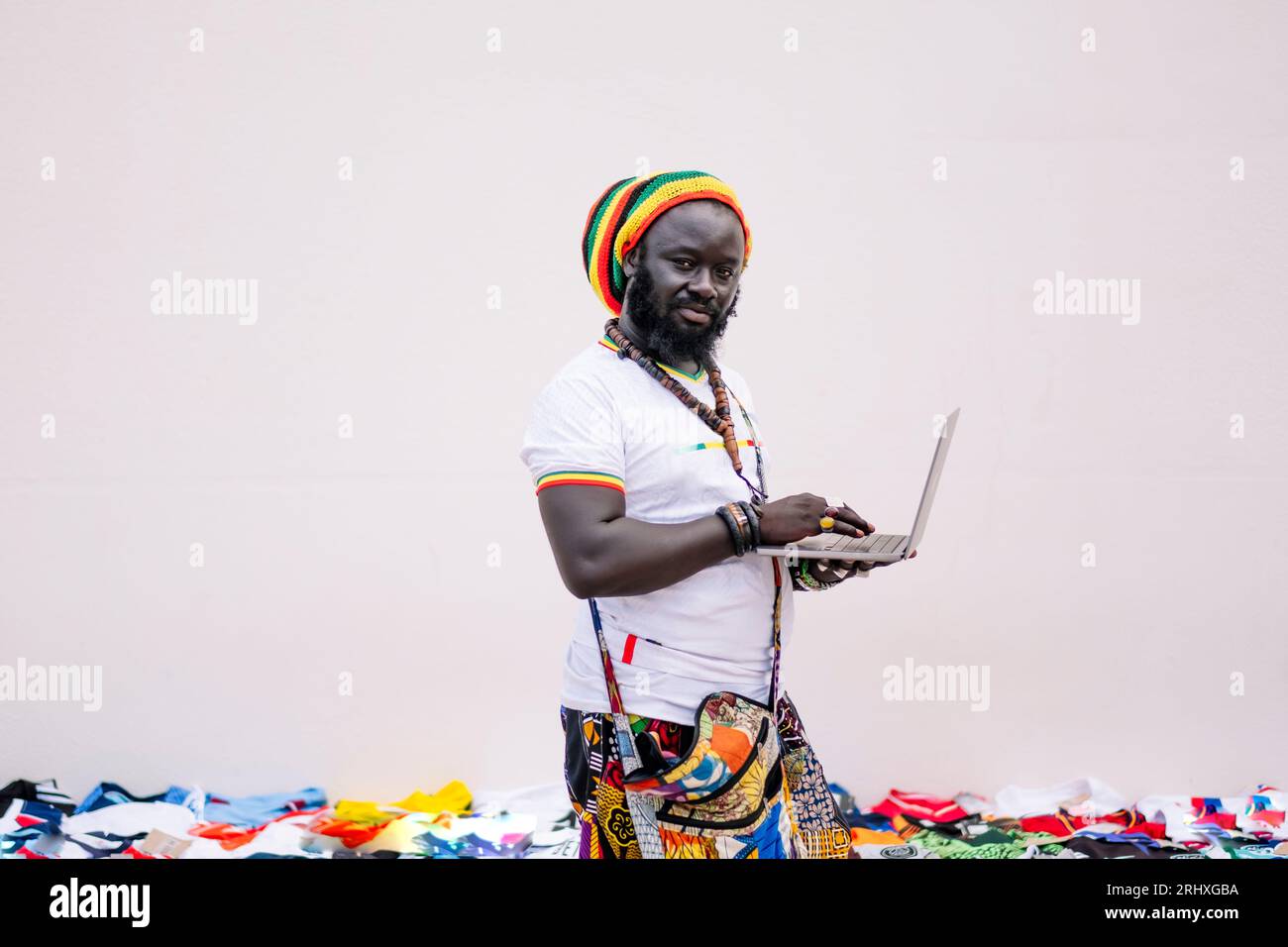 Vue latérale de l'homme afro-américain dans des vêtements colorés et chapeau debout et regardant la caméra tout en tapant sur l'ordinateur portable sur fond blanc avec dif Banque D'Images