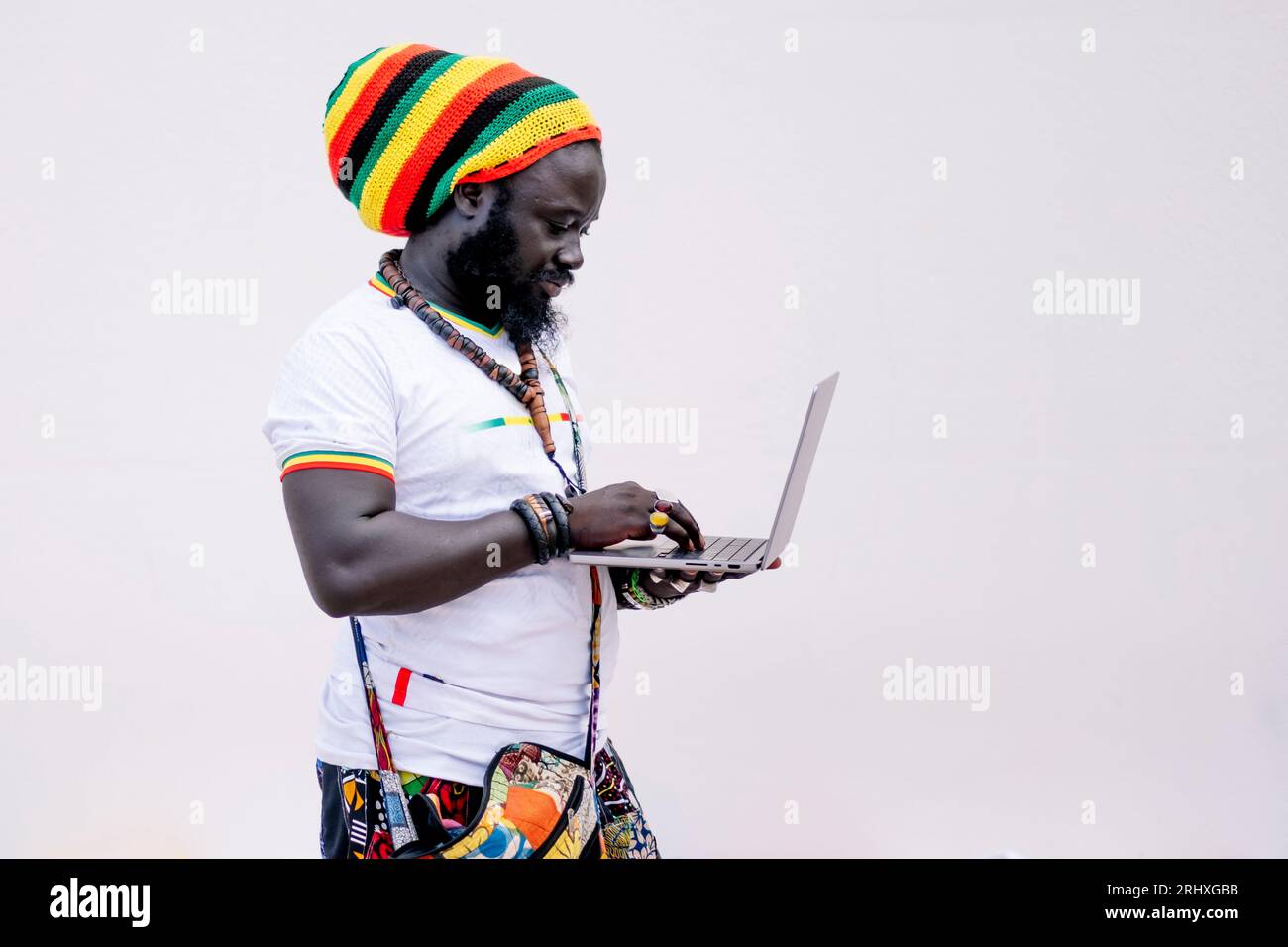 Vue latérale de l'homme afro-américain dans les vêtements colorés et chapeau debout et regardant l'écran tout en tapant sur l'ordinateur portable sur fond blanc Banque D'Images
