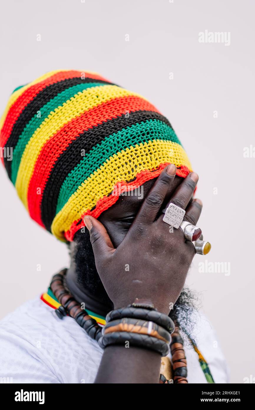 Homme afro-américain anonyme dans le chapeau coloré debout et regardant loin tout en couvrant le visage avec la main sur fond blanc Banque D'Images