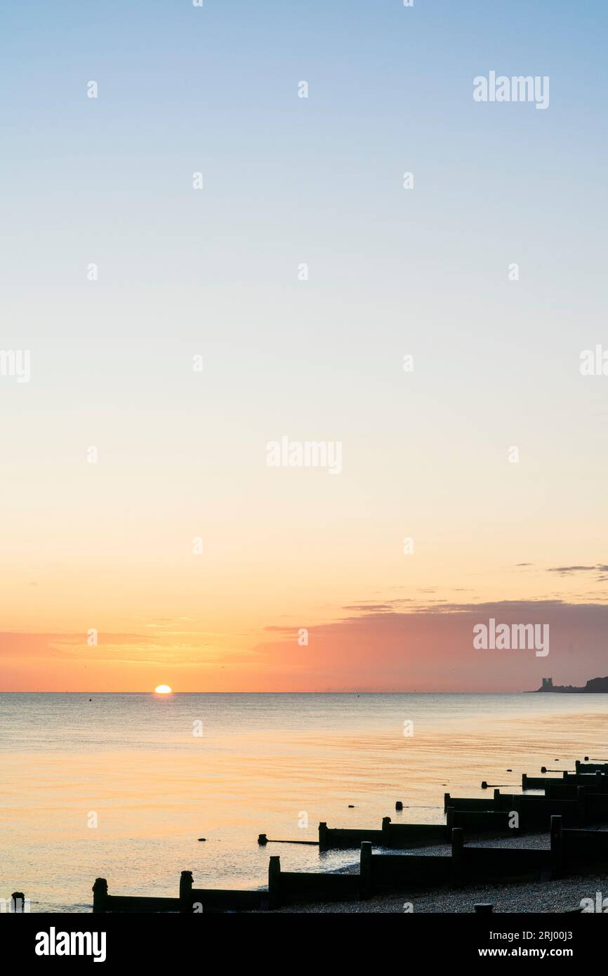 Lever de soleil sur la mer vu de la plage avec des brise-vagues en bois, alias groynes, à Herne Bay sur la côte nord du Kent. Un nuage à l'horizon, sinon un ciel dégagé. Mer très calme. Banque D'Images