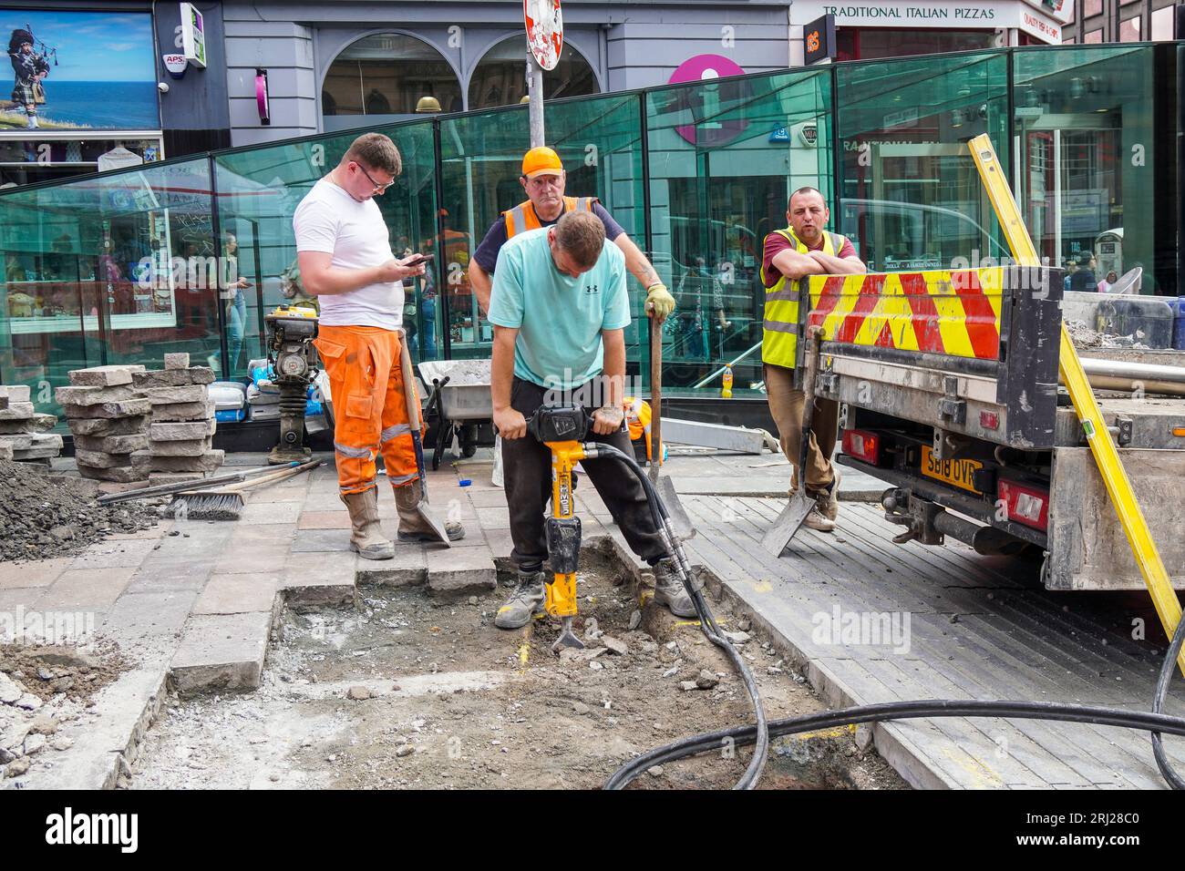 4 ouvriers réparant la chaussée, dont un travaillant sur une perceuse routière, un appuyé sur le camion, un sur son téléphone et un appuyé sur sa pelle, Glasgow, Banque D'Images