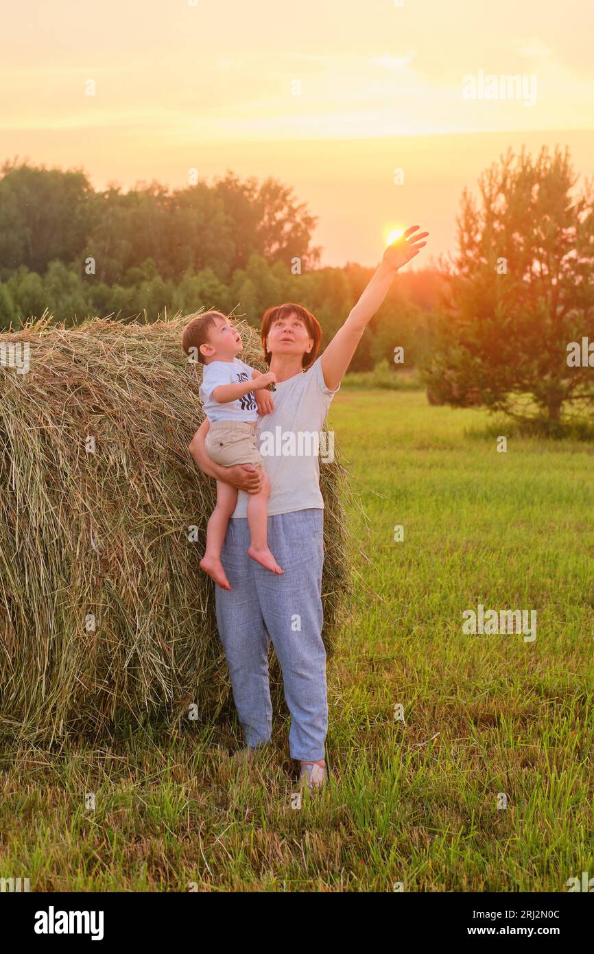Le soleil se couche sur la ferme comme une femme âgée et bébé sourit devant une pile de foin. Portrait d'un enfant heureux âgé de deux ans et d'une femme aînée de si Banque D'Images