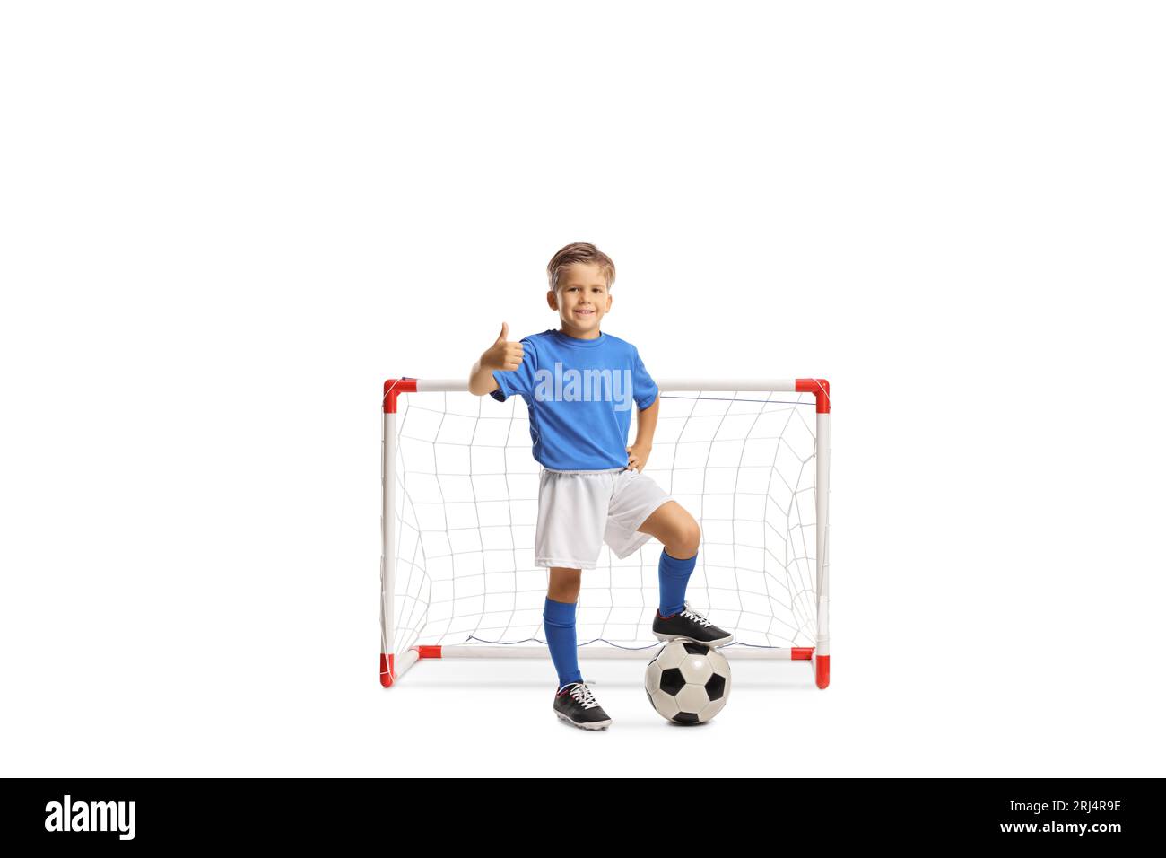 Garçon dans un maillot de sport marchant sur un ballon de football et faisant des gestes pouces vers le haut devant un mini but isolé sur fond blanc Banque D'Images