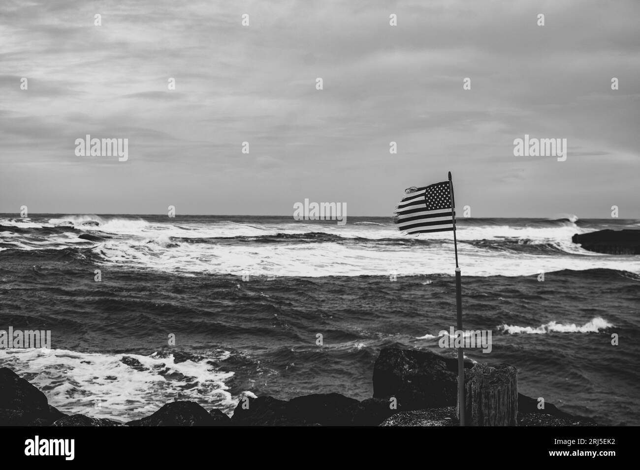 Une échelle de gris de drapeau américain agitant sur la plage contre les vagues de l'océan à Bandon, Oregon Banque D'Images