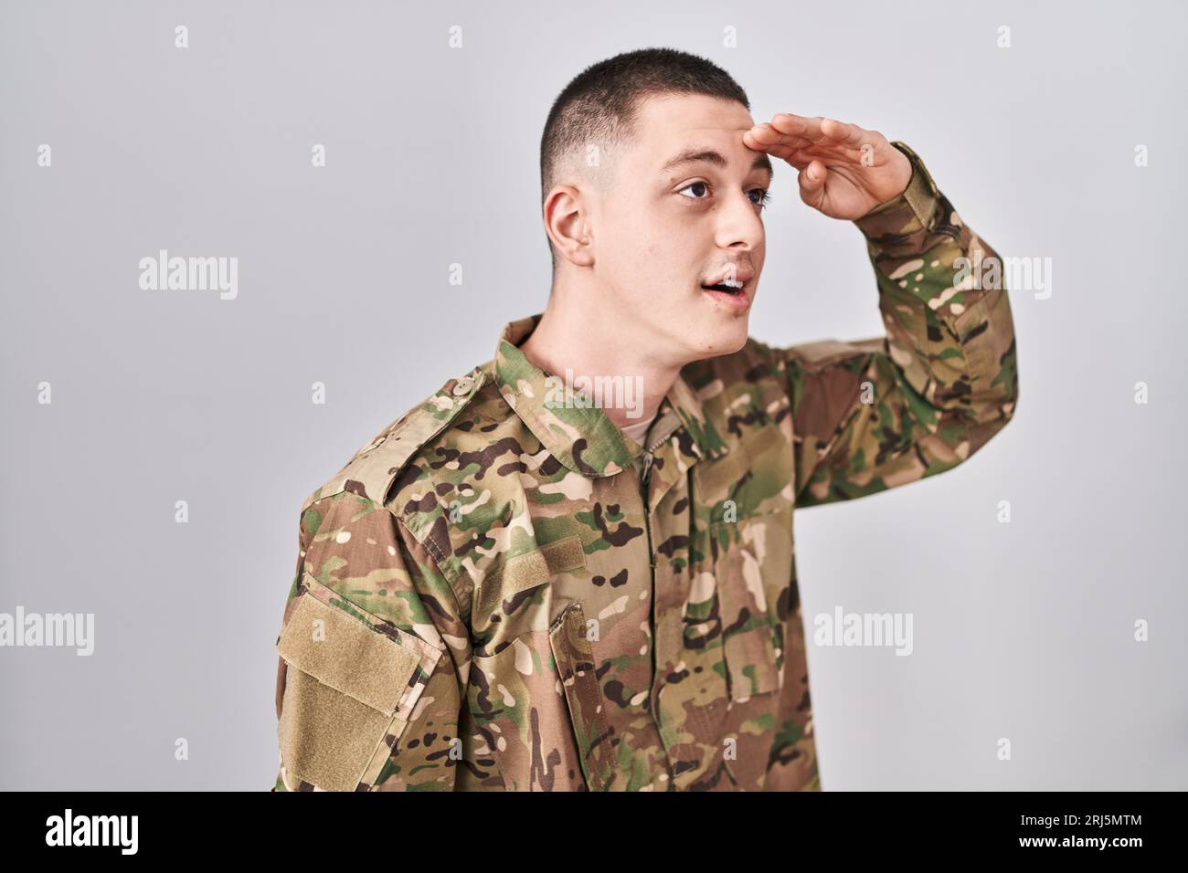 Jeune homme portant l'uniforme militaire de camouflage très heureux et souriant regardant loin avec la main sur la tête. concept de recherche. Banque D'Images