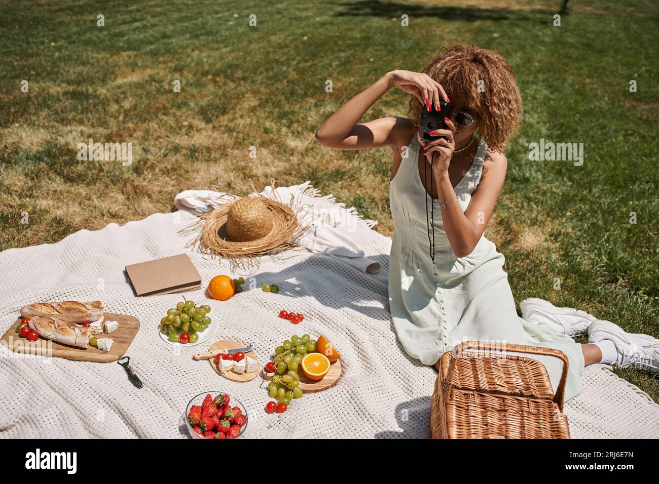 femme afro-américaine prenant la photo sur l'appareil photo vintage près des fruits et légumes, pique-nique d'été Banque D'Images