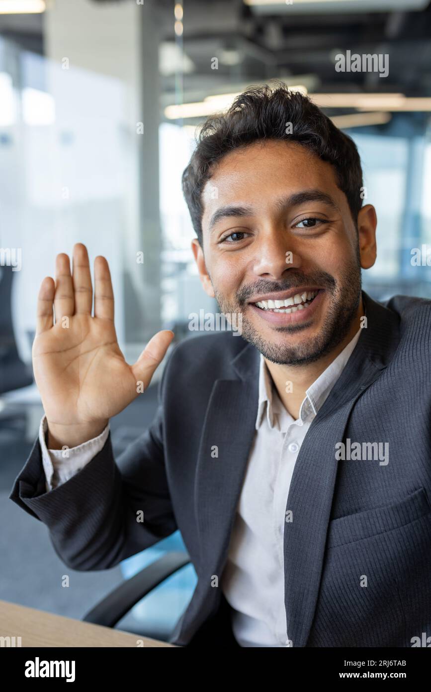Photo verticale, jeune homme d'affaires prospère regardant dans la caméra smartphone parler sur appel vidéo en utilisant l'application sur le téléphone, souriant et ayant une conversation amicale avec des amis et des collègues à l'intérieur du bureau. Banque D'Images
