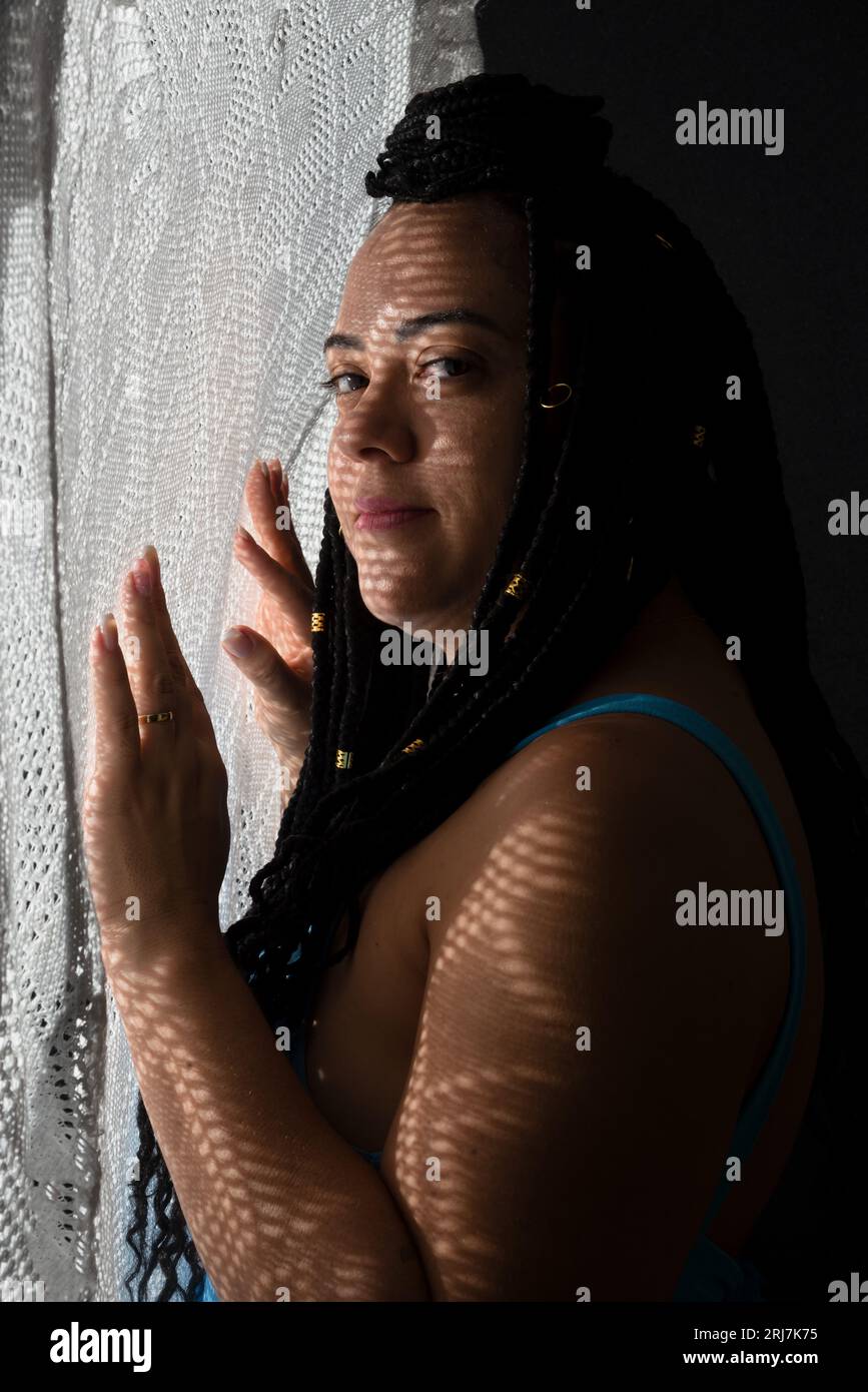 Photo d'une belle femme avec des tresses dans les cheveux et des ombres géométriques sur son visage avec ses mains sur un tissu blanc. Portrait de studio avec fond noir Banque D'Images