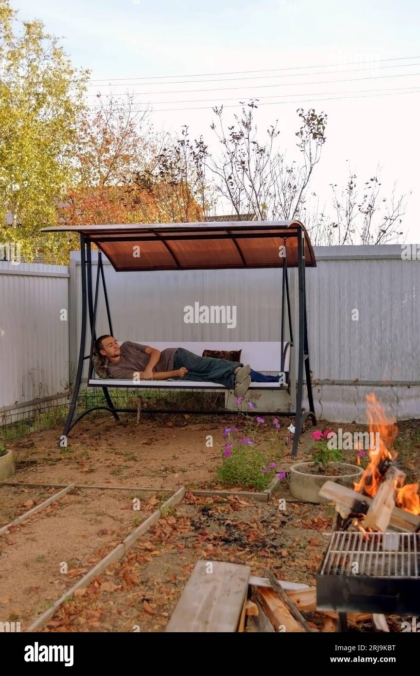 Jeune homme endormi allongé sur une balançoire de jardin à côté d'un feu de joie Banque D'Images