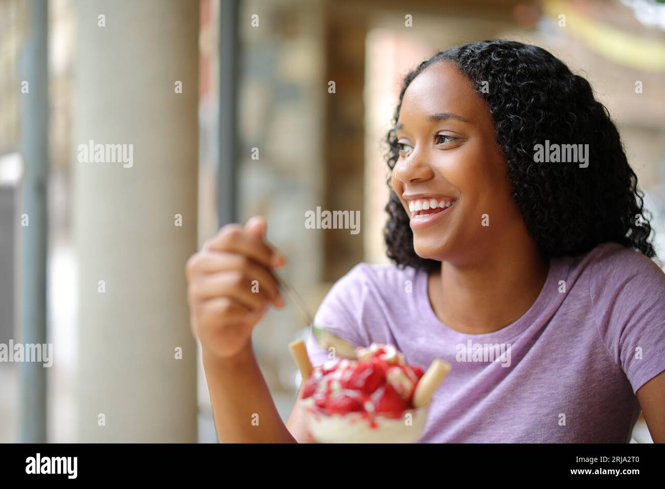 Heureuse femme noire mangeant le dessert regardant loin dans une terrasse de café Banque D'Images