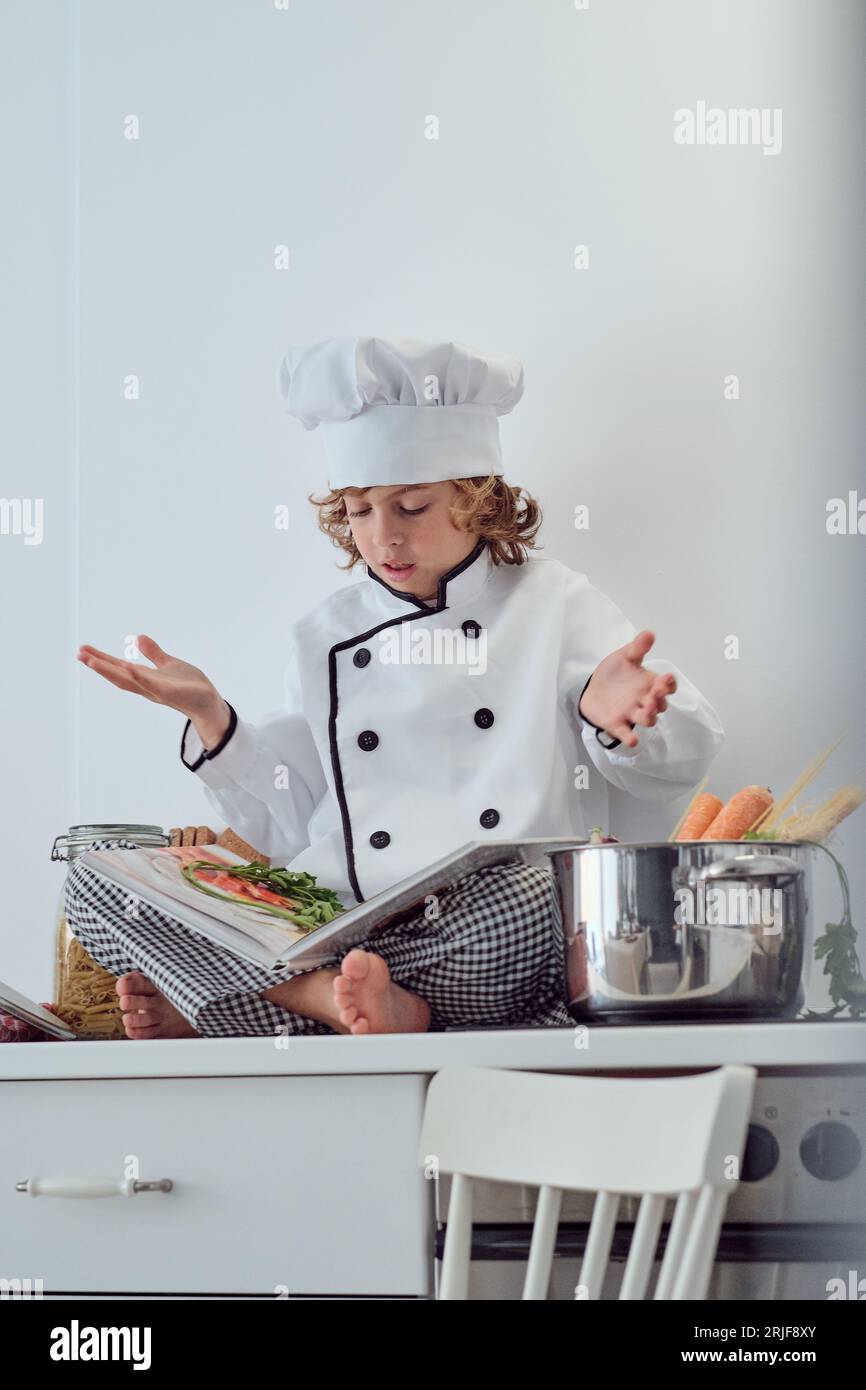 De dessous de garçon préadolescent plein corps en costume de chef et chapeau assis sur le comptoir de cuisine et lisant le livre culinaire et exprimant la confusion de la recette Banque D'Images