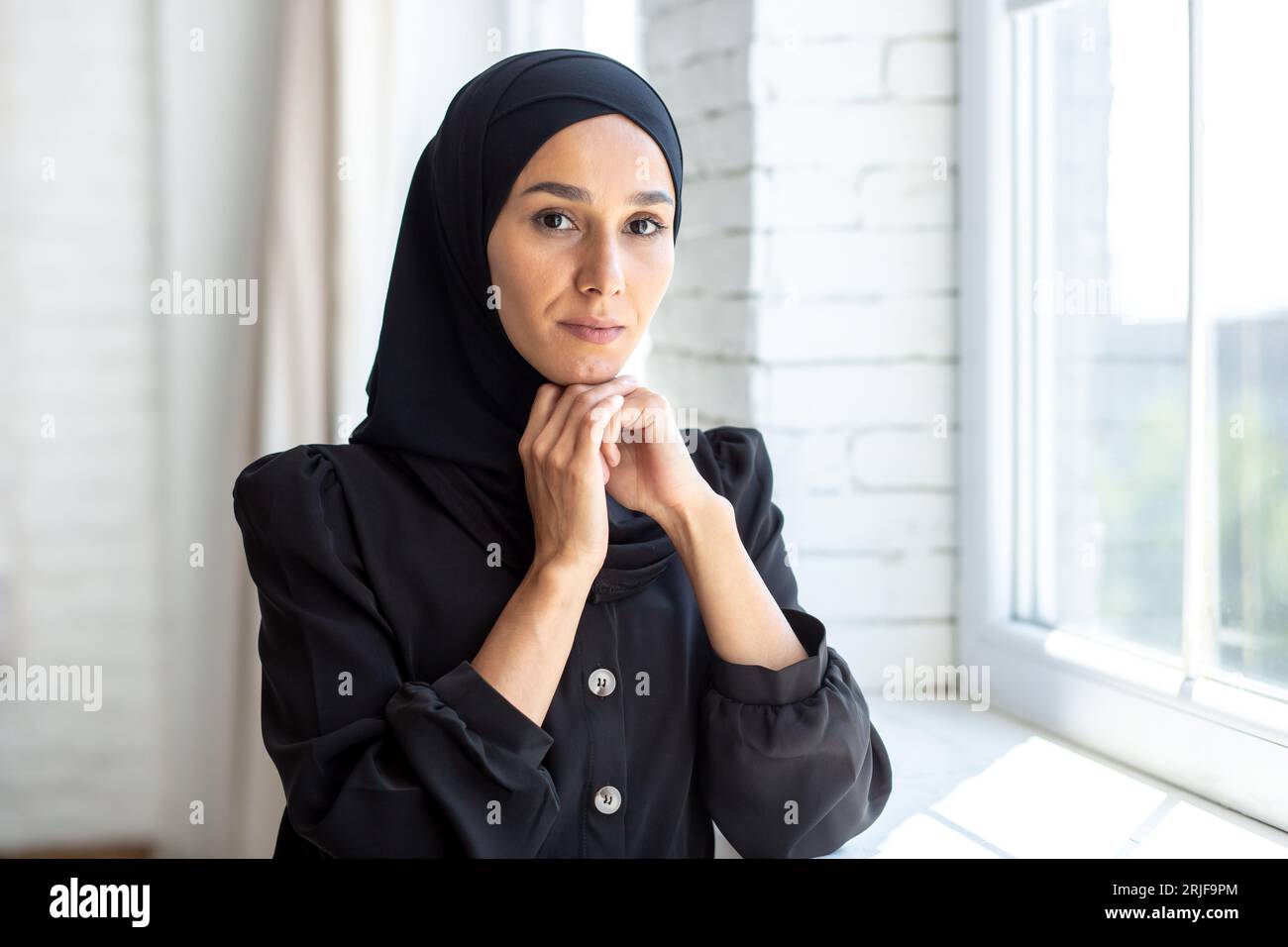 Portrait d'une jeune femme musulmane dans un hijab noir debout à l'intérieur près de la fenêtre, pliant ses bras et posant et regardant sérieusement la caméra. Banque D'Images