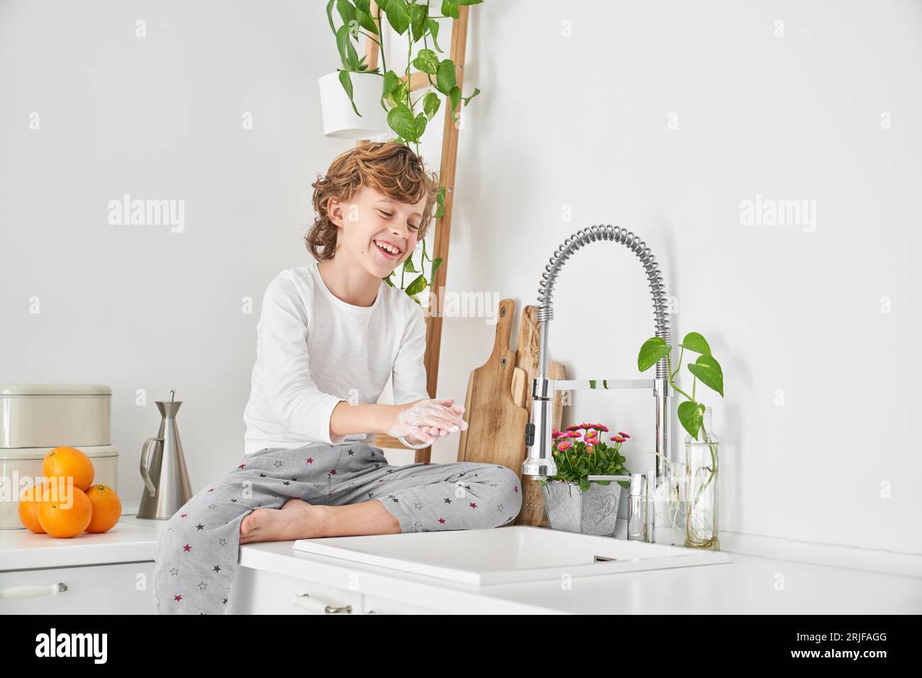 Garçon aux cheveux bouclés pieds nus assis près de l'évier et se lavant les mains avec du savon pour prévenir la propagation des bactéries dans la cuisine le matin Banque D'Images