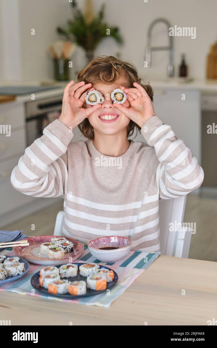 Enfant préadolescent souriant et mettant des sushis sur les yeux tout en étant assis à table contre l'assiette avec un ensemble de rouleaux Banque D'Images