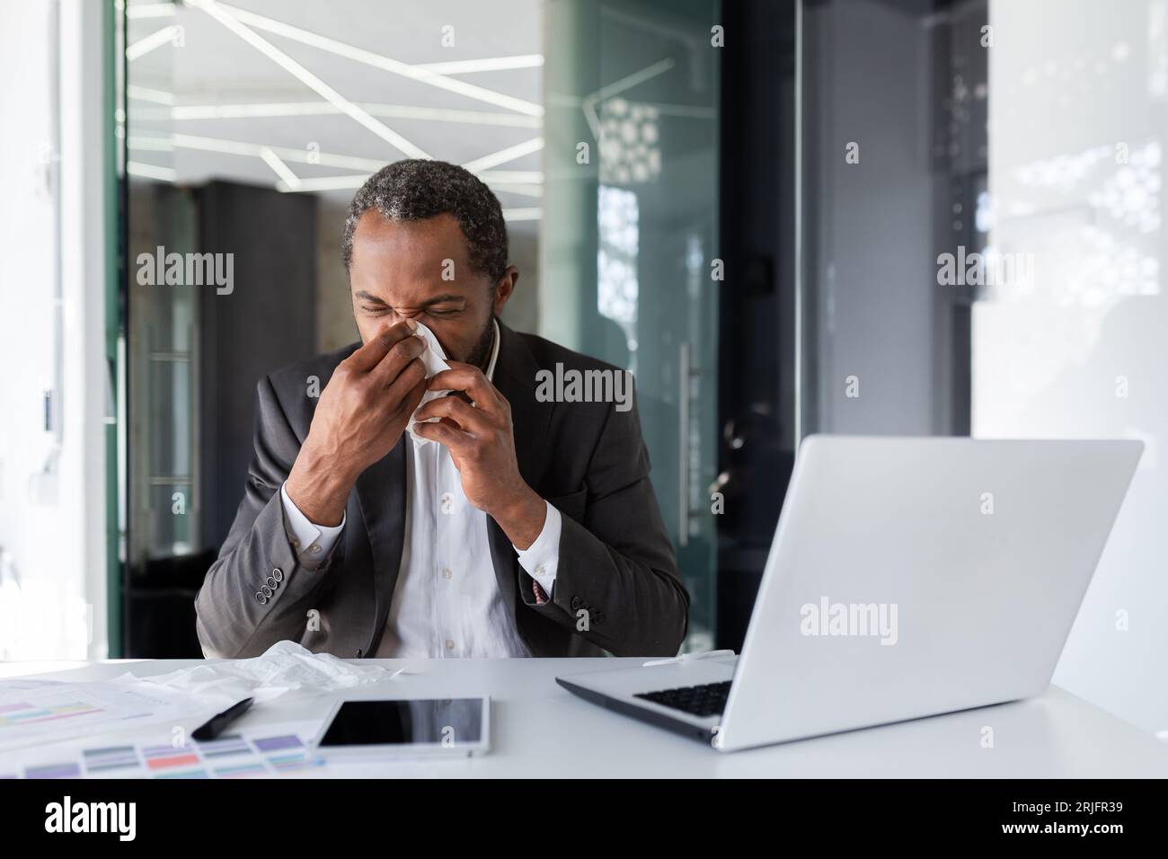 Homme malade au lieu de travail à l'intérieur du bureau avec joie et froid, homme d'affaires éternue assis au bureau, homme afro-américain avec froid utilise un ordinateur portable au travail. Banque D'Images