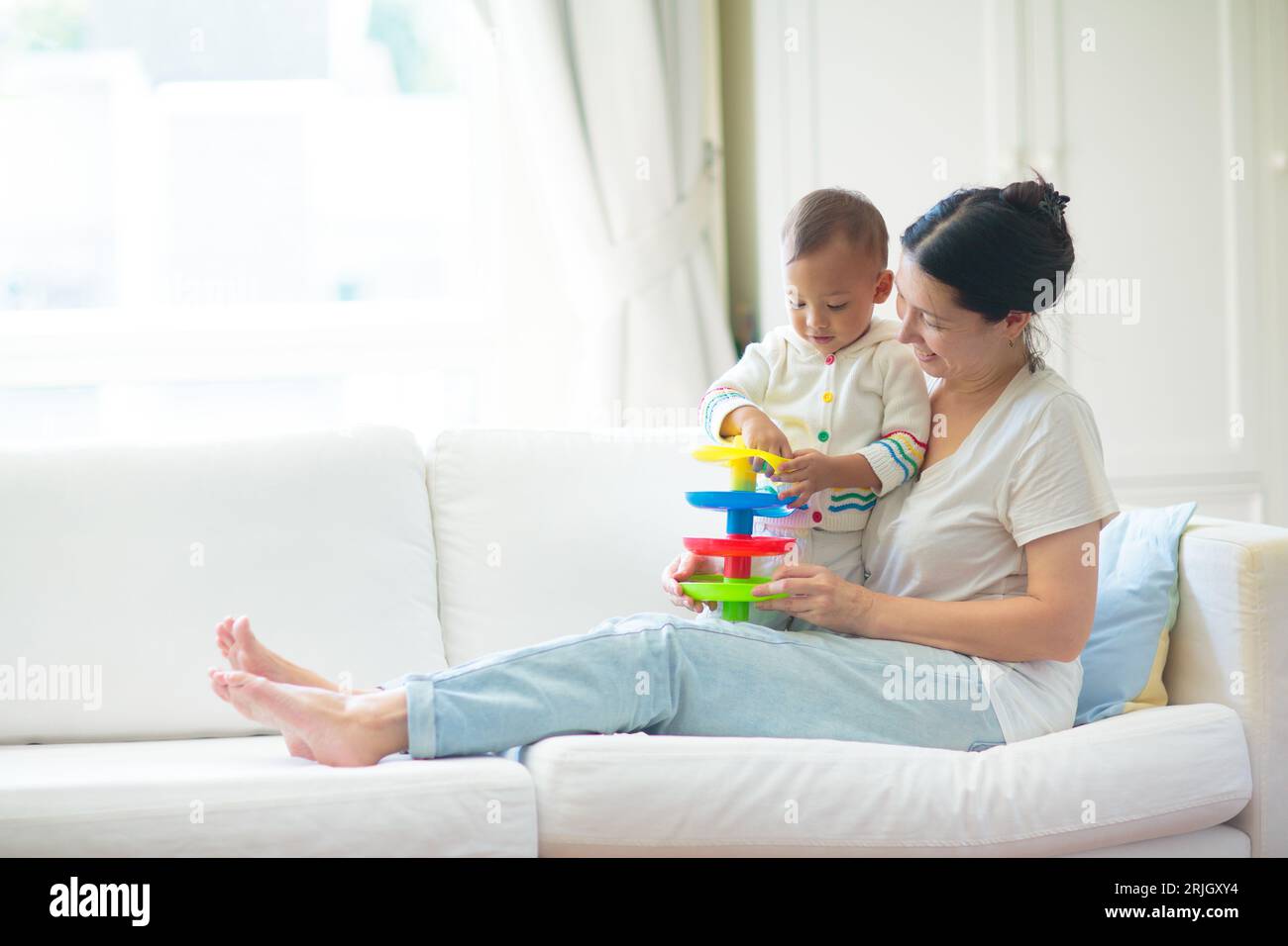 Mère et enfant sur un canapé blanc. maman asiatique et bébé garçon jouant dans la chambre ensoleillée. Parent et petit enfant se relaxant à la maison. Banque D'Images