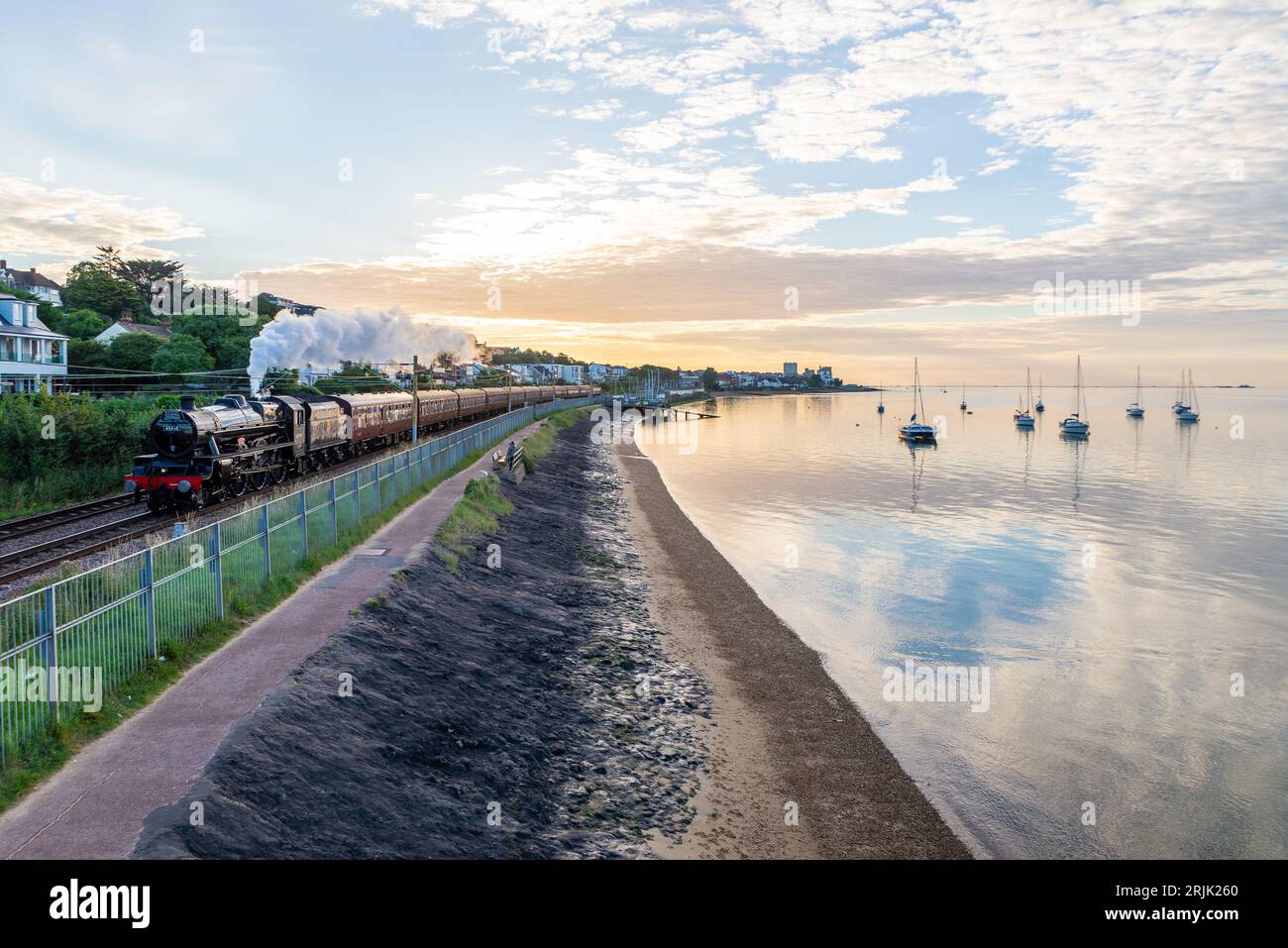 Chalkwell, Southend on Sea, Essex, Royaume-Uni. 23 août 2023. Un train à vapeur a quitté Southend on Sea tôt le matin en direction de Bristol Temple Meads, en passant le long de l'estuaire calme et plat de la Tamise à Chalkwell. Le train spécial organisé par la Railway Touring Company est transporté par l'ancienne locomotive à vapeur LMS Jubilee Class 45690 Leander. Leander est l'une des locomotives à vapeur autorisées à circuler sur les lignes principales pour des voyages spéciaux et a été construite en 1936. Il a été renuméroté 45690 par British Railways en 1948 à la suite de la nationalisation. Yachts sur l'eau de l'estuaire qui reflète la lumière de l'aube Banque D'Images