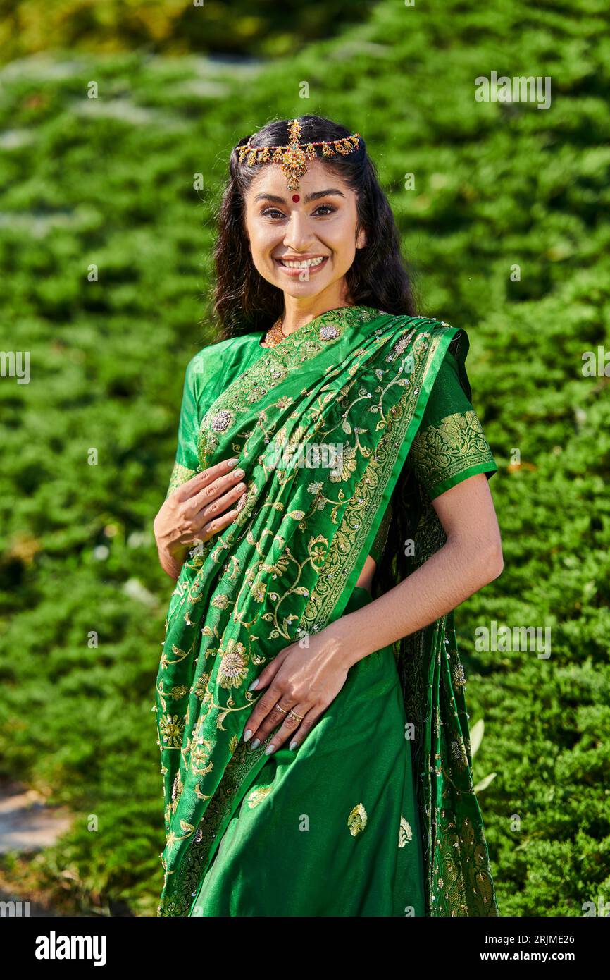 portrait de jeune femme indienne joyeuse dans sari traditionnel et matha patti regardant la caméra dans le parc Banque D'Images